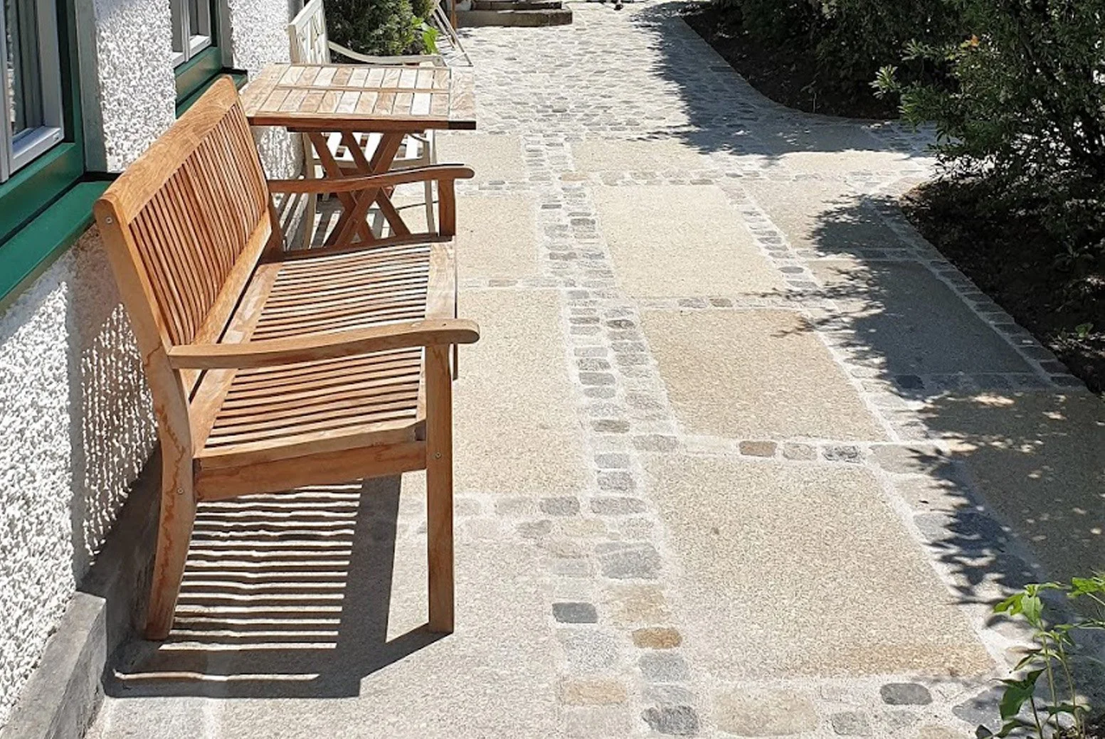 Holzbank und kleiner Tisch auf einem gepflasterten Weg neben einem Haus mit weißen Wänden und grünen Fenstern, im Schatten von Bäumen.