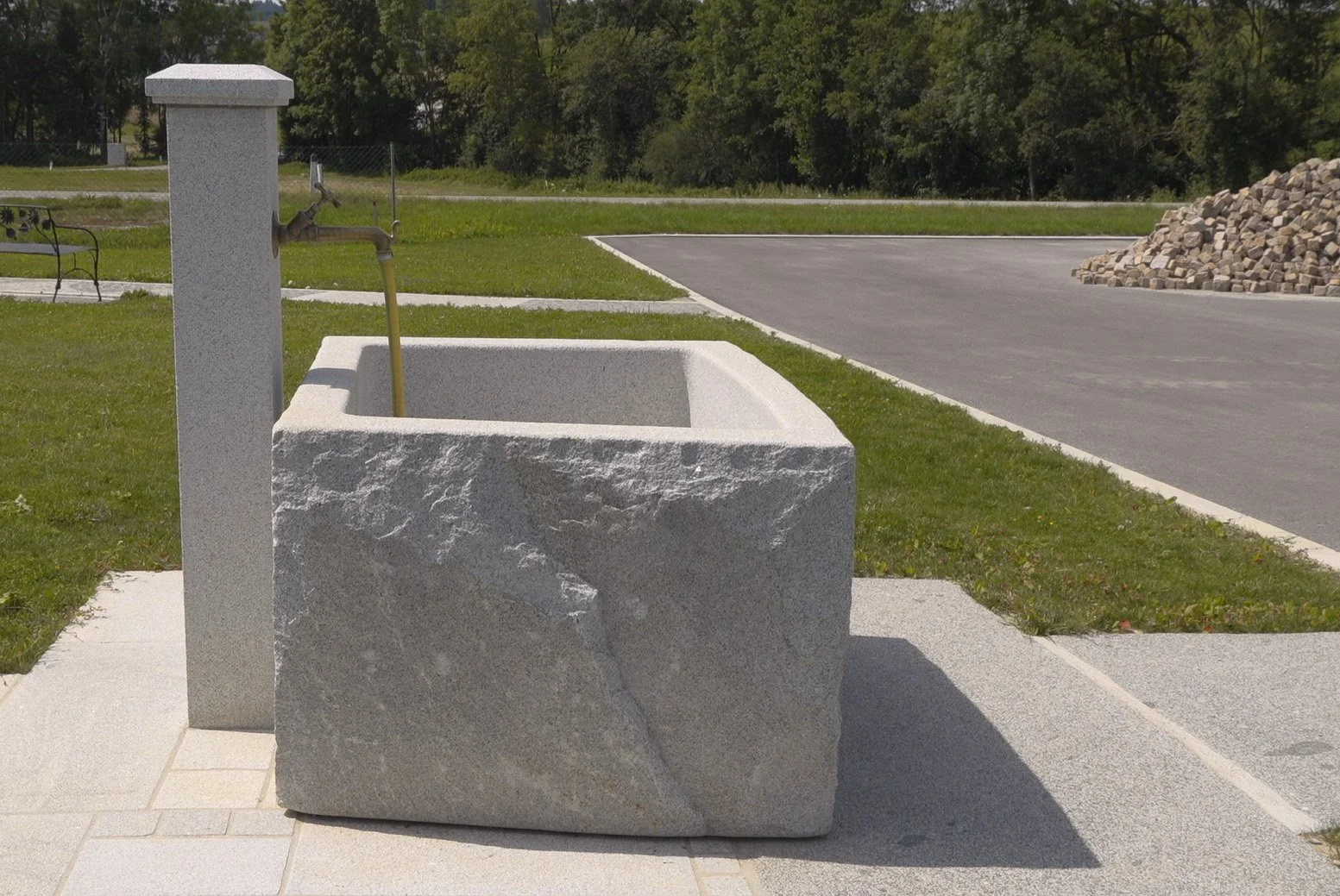Steinwaschbecken mit Wasserspender, im Freien auf gepflastertem Boden neben einer Rasenfläche und Straße.