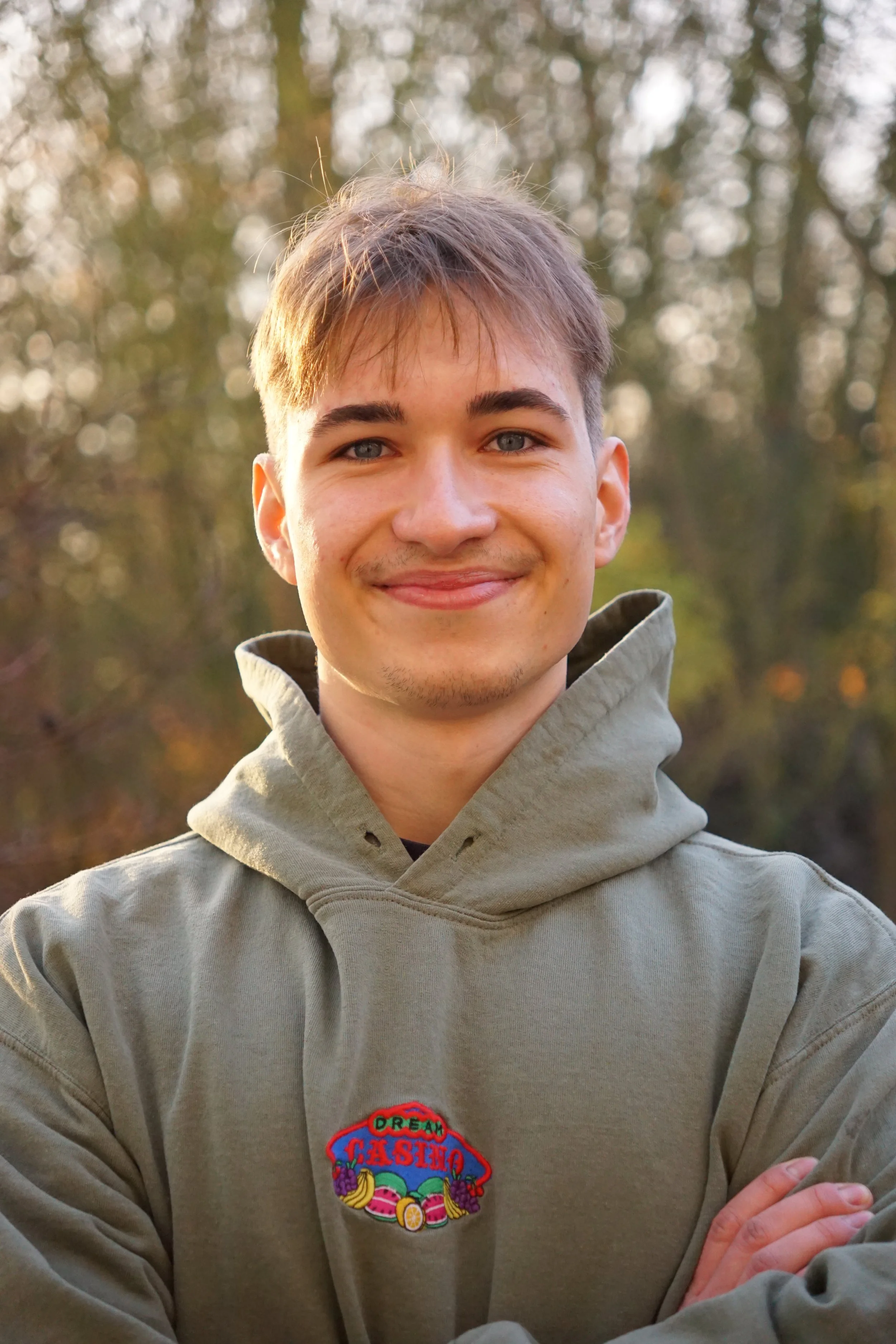 Ein lachender junger Mann trägt einen grauen Hoodie mit einem bunten Aufnäher und steht vor einem Hintergrund aus Bäumen im Freien.