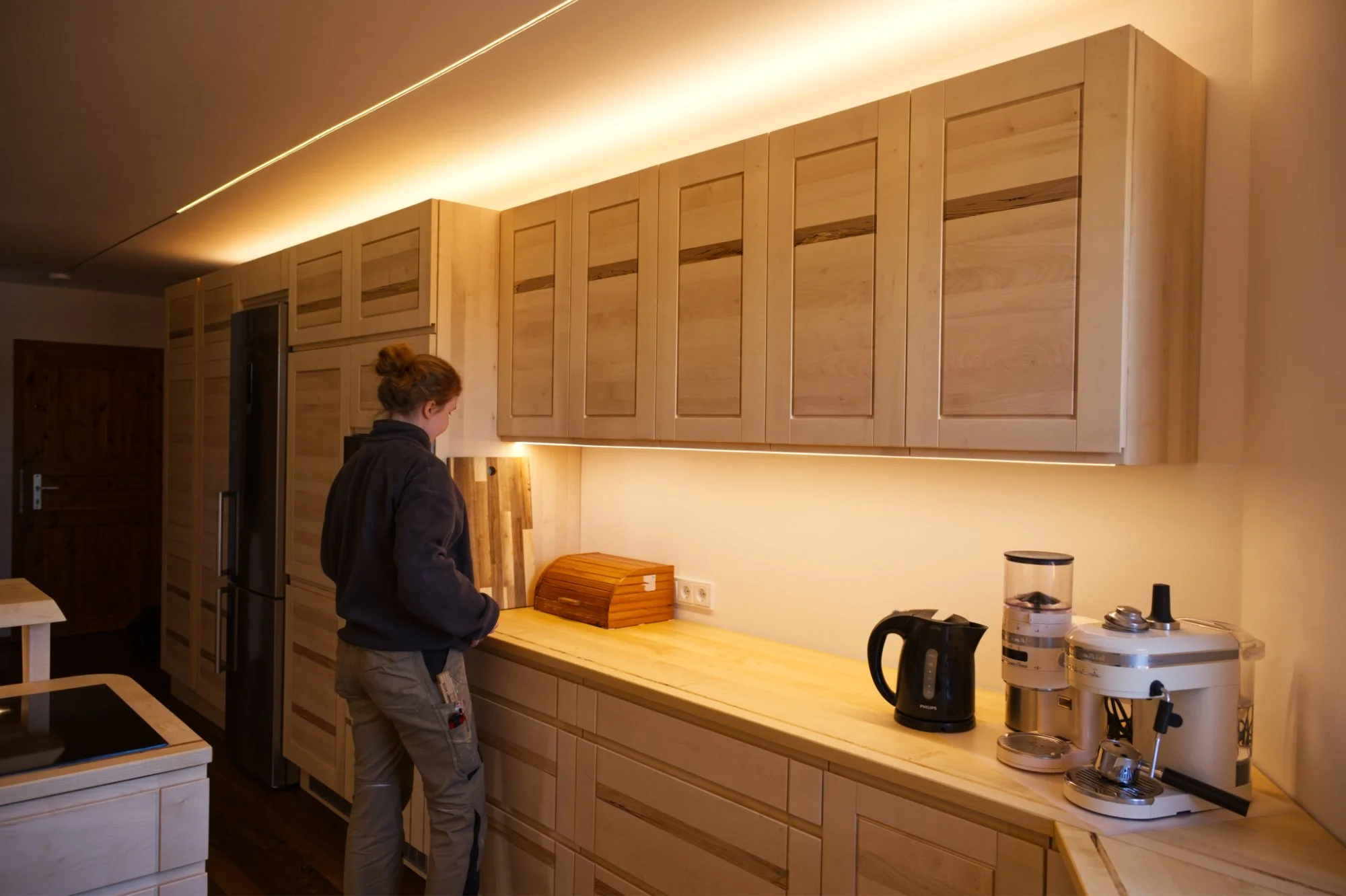 Eine Person steht in einer modernen, hellen Küche mit hellen Holzmöbeln, während sie an der Arbeitsplatte arbeitet, auf der Küchenutensilien und ein Brotkasten stehen.