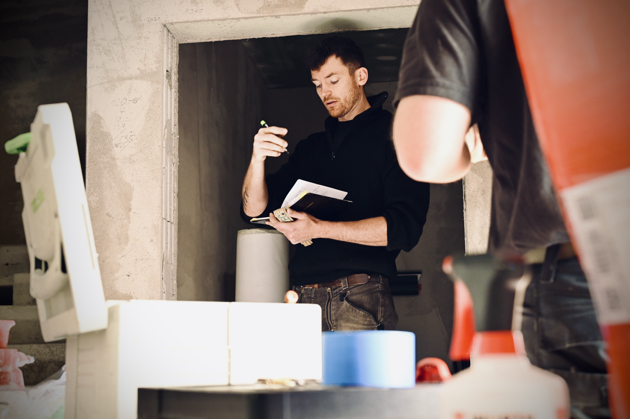 Mann steht in einer Baustelle, hält einen Notizblock und einen Stift und liest oder schreibt, während eine zweite Person anwesend ist.