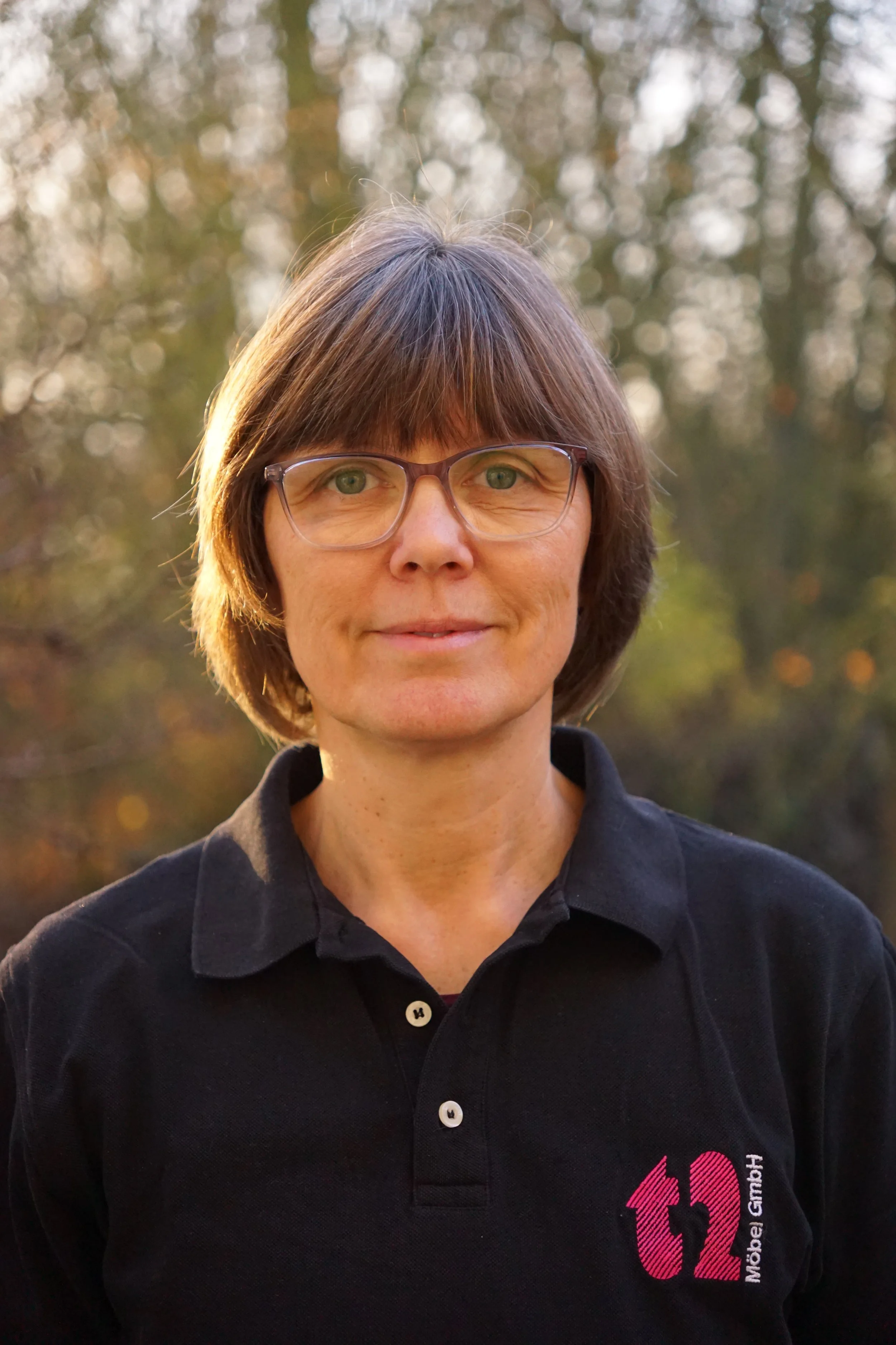 Porträt einer Frau mit Brille, kurze braune Haare, schwarzem Polo-Shirt mit Logo 't2 Möbel GmbH', vor unscharfem Wald im Hintergrund.