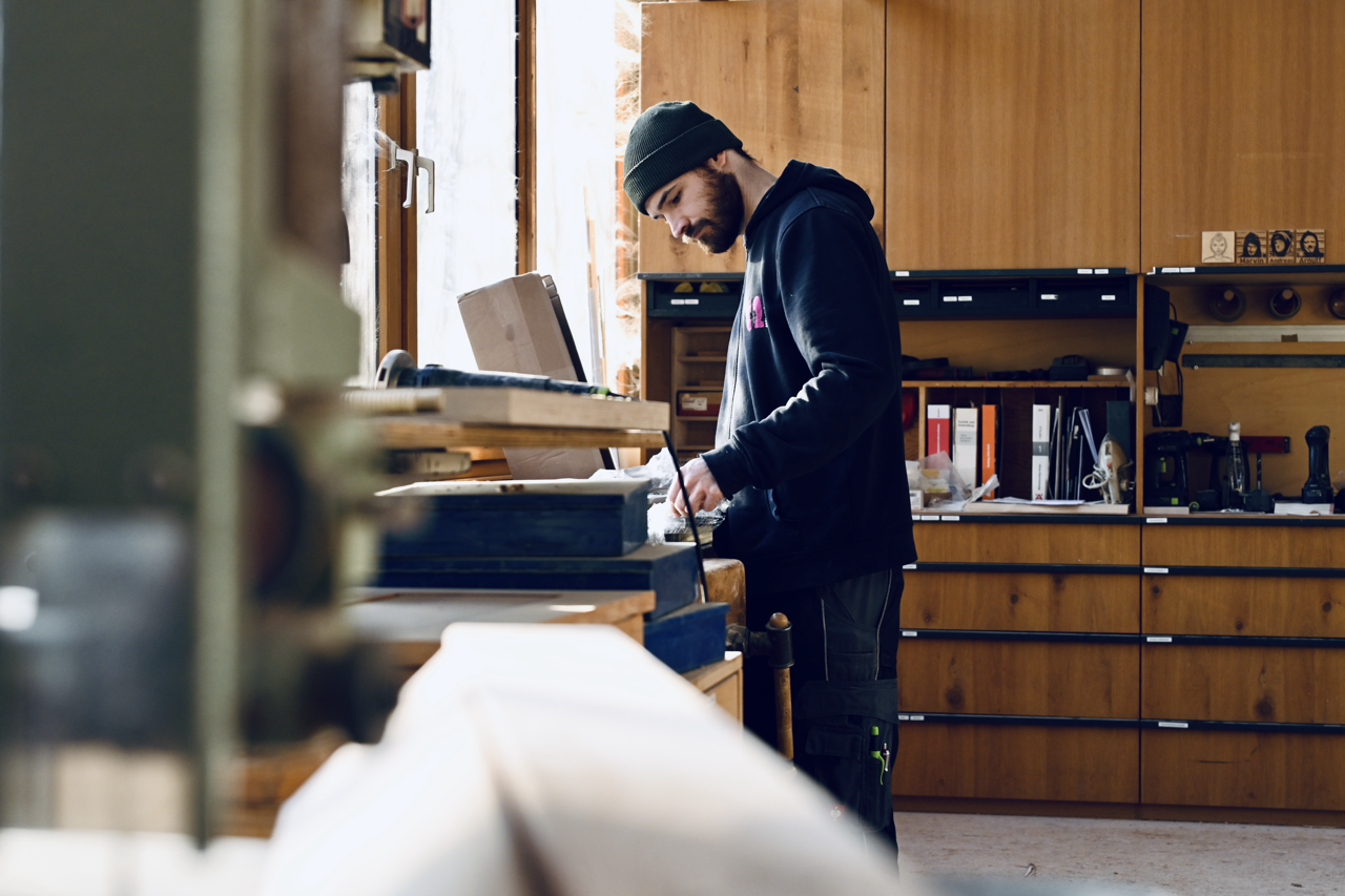 Ein Mann arbeitet in einer Holzwerkstatt an einer werkbank, umgeben von Werkzeugen und Holzmöbeln.