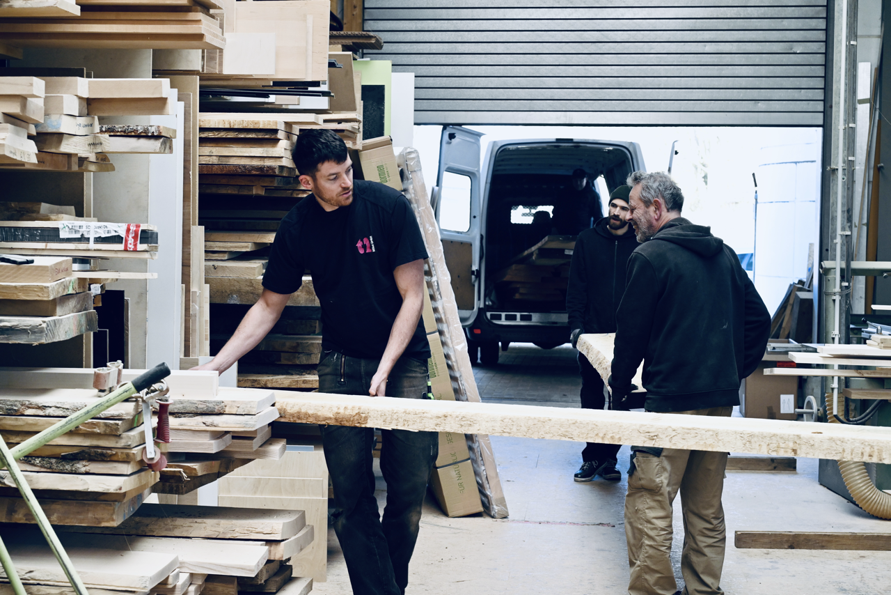 Drei Männer arbeiten in einer Holzwerkstatt, einige Holzplatten sind gestapelt, und ein Mann hält ein großes Brett. Im Hintergrund ein LKW mit geöffnetem Hintertor.