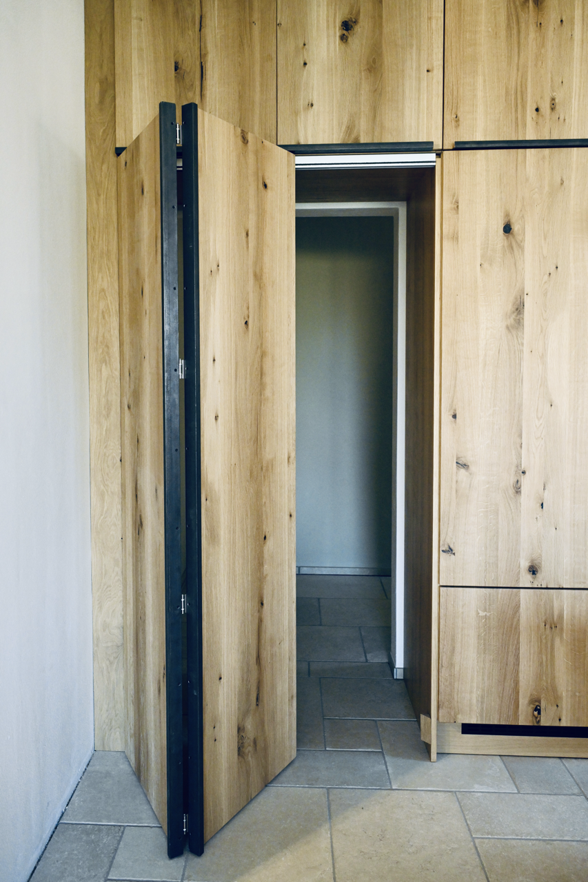 Offene Holzklapplade, teilweise geöffnet, in einer Holzverkleidung, neben Fliesenboden.