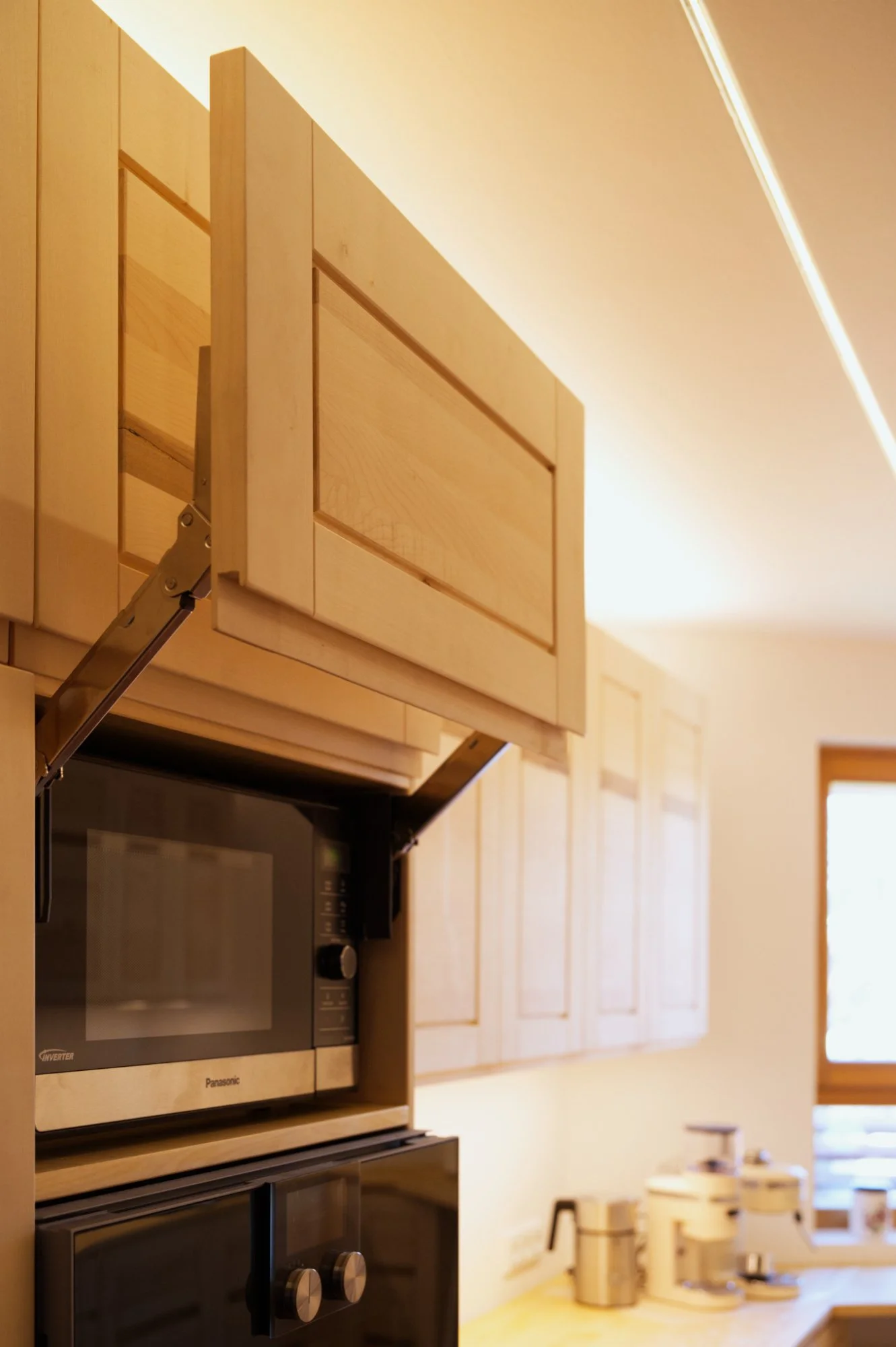 Küche mit massivholz Schränken, Mikrowelle und Kaffeemaschine, Blick auf eine Fensterfront.