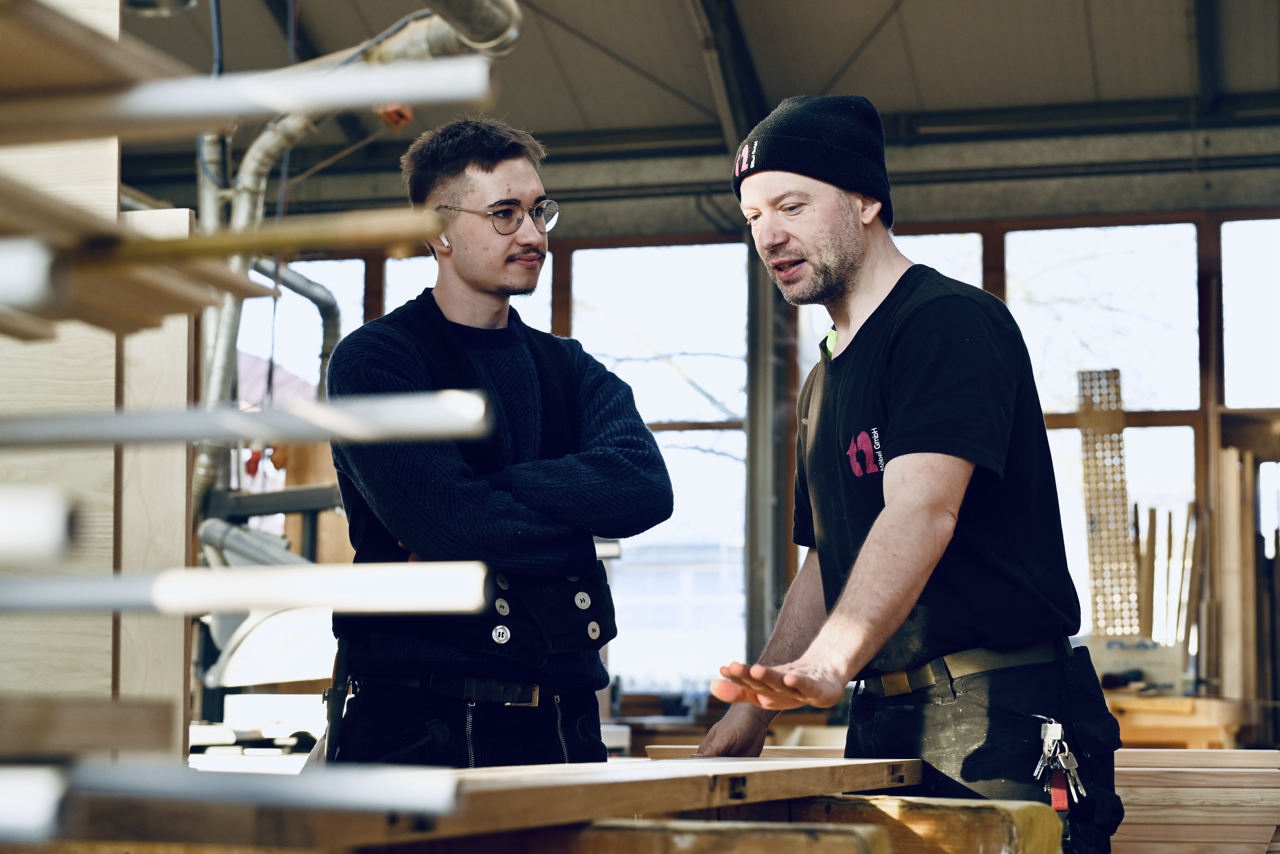 Zwei Männer sprechen in einer Werkstatt, einer erklärt dem anderen etwas, während sie an einem Holzstück arbeiten.