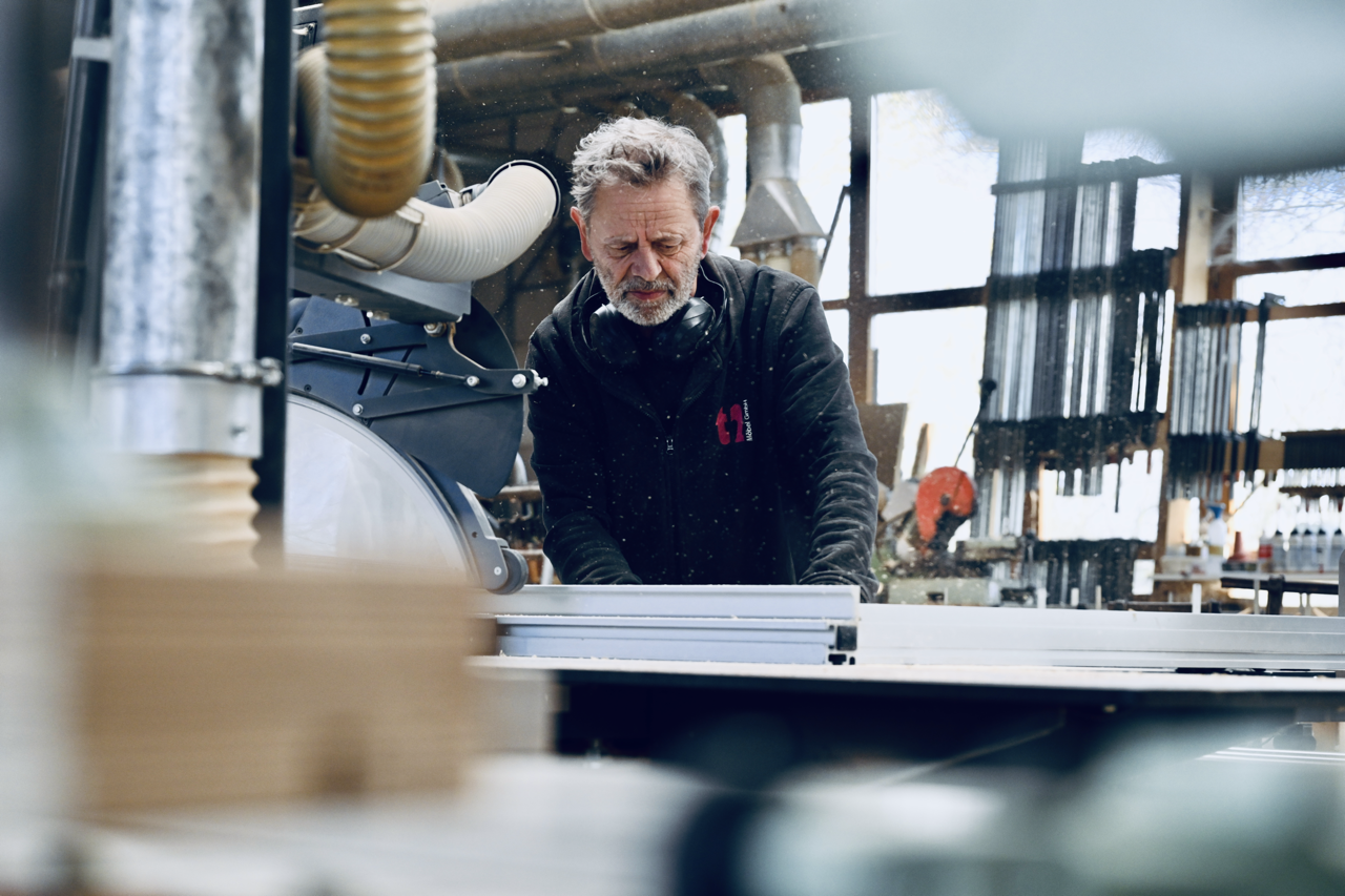 Mann arbeitet in einer Holzwerkstatt mit einer Kreissäge, trägt Schutzbrille und Kopfhörer, um sich vor Staub und Lärm zu schützen.