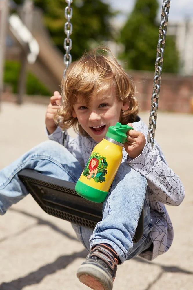 Ein Junge sitzt auf einer Schaukel im Freien und hält eine bunte Wasserflasche mit Löwen-Design in der Hand.