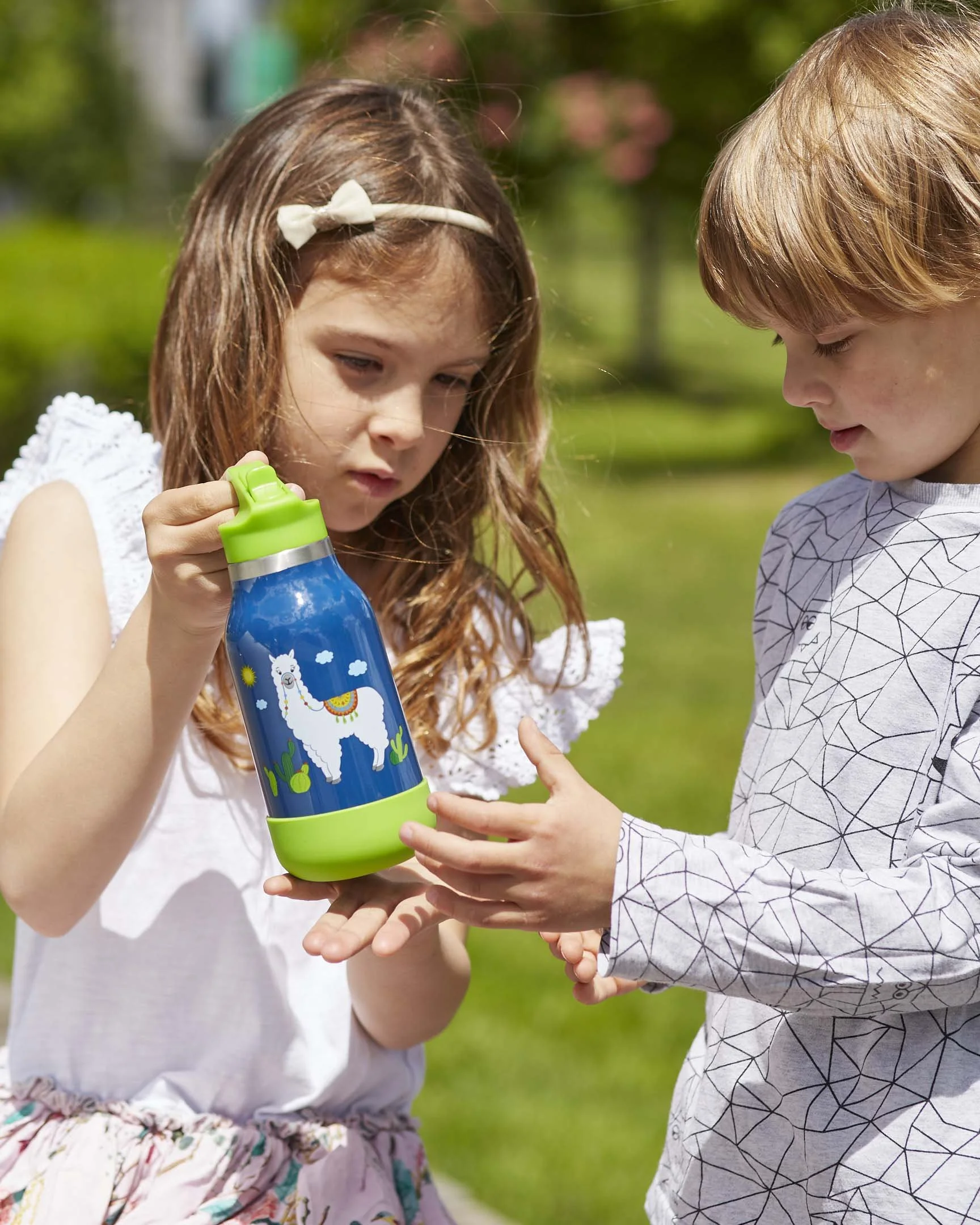 Zwei Kinder, ein Mädchen mit braunen Haaren und ein Junge mit blonden Haaren, schauen auf eine Wasserflasche in der Hand des Mädchens, die eine Lama-Illustration hat, draußen im Grünen bei Sonnenschein.