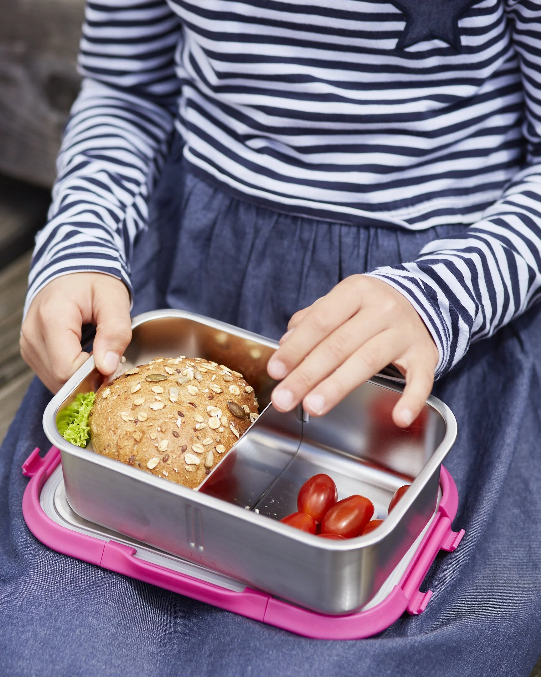 Ein Mädchen mit gestreiftem Kleid hält eine Brotdose mit einem Sesambrötchen, Tomaten und Salat darin.