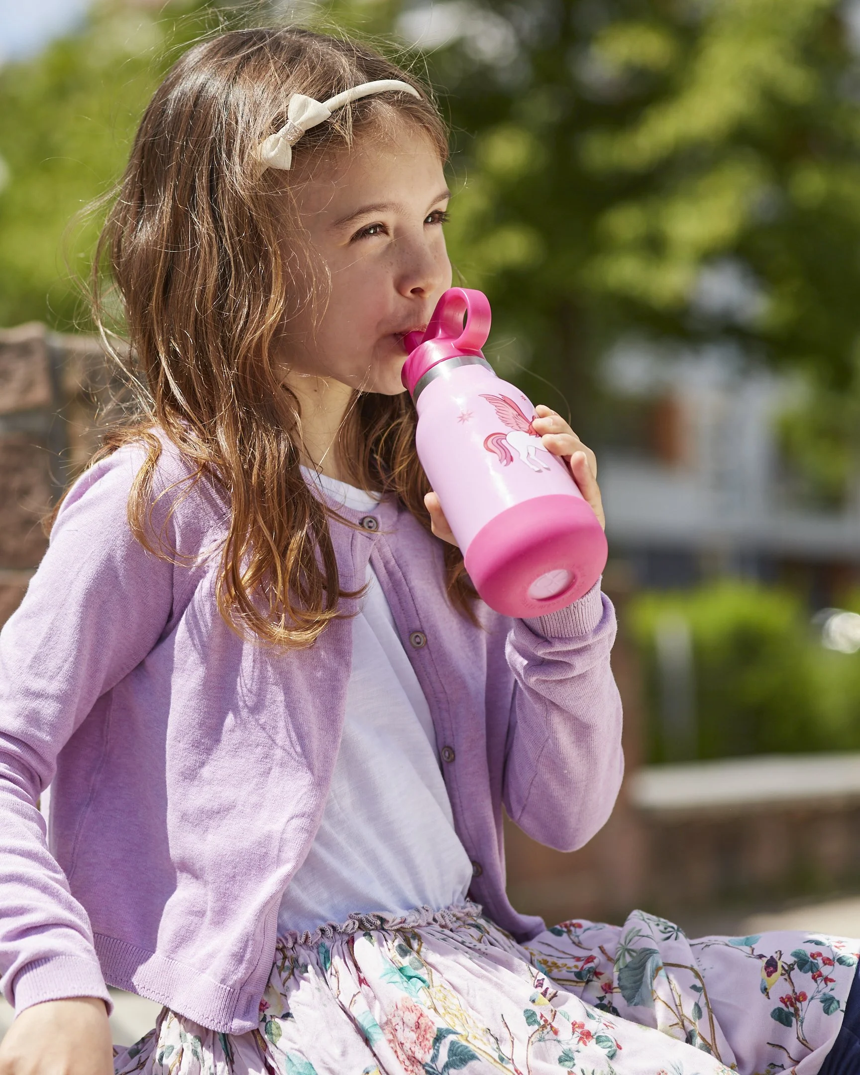 Ein junges Mädchen mit lang, lockigem Haar trinkt aus einer pinken Trinkflasche mit Einhorn-Design im Freien an einem sonnigen Tag.