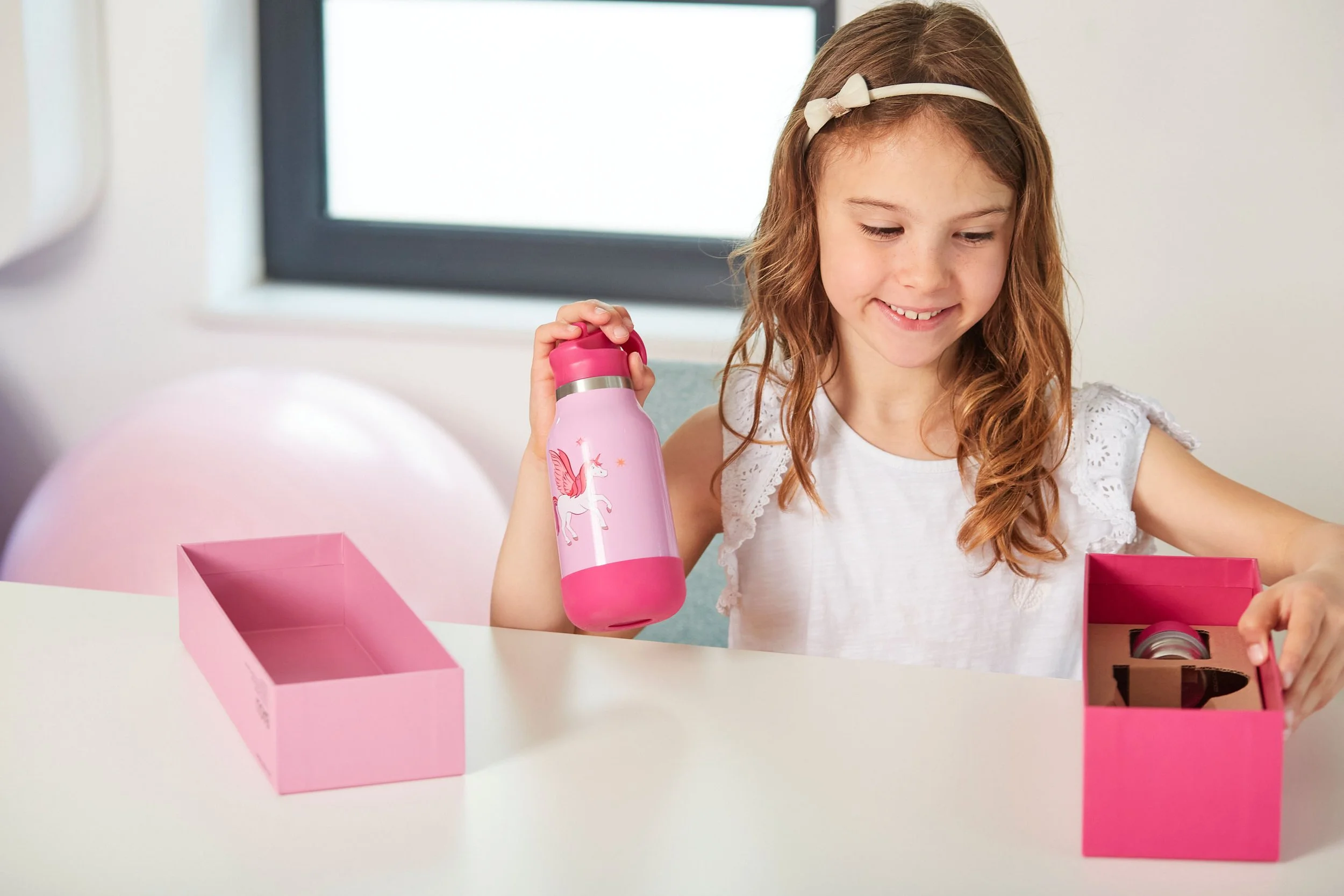 Ein lachendes Mädchen mit Schulterlangem, lockigem, braunem Haar, hält eine pinke Wasserflasche mit Einhorn-Design. Sie sitzt an einem weißen Tisch, auf dem ein leerer rosa Karton und eine weitere Wasserflasche in einem rosa Karton liegen. Im Hinterg