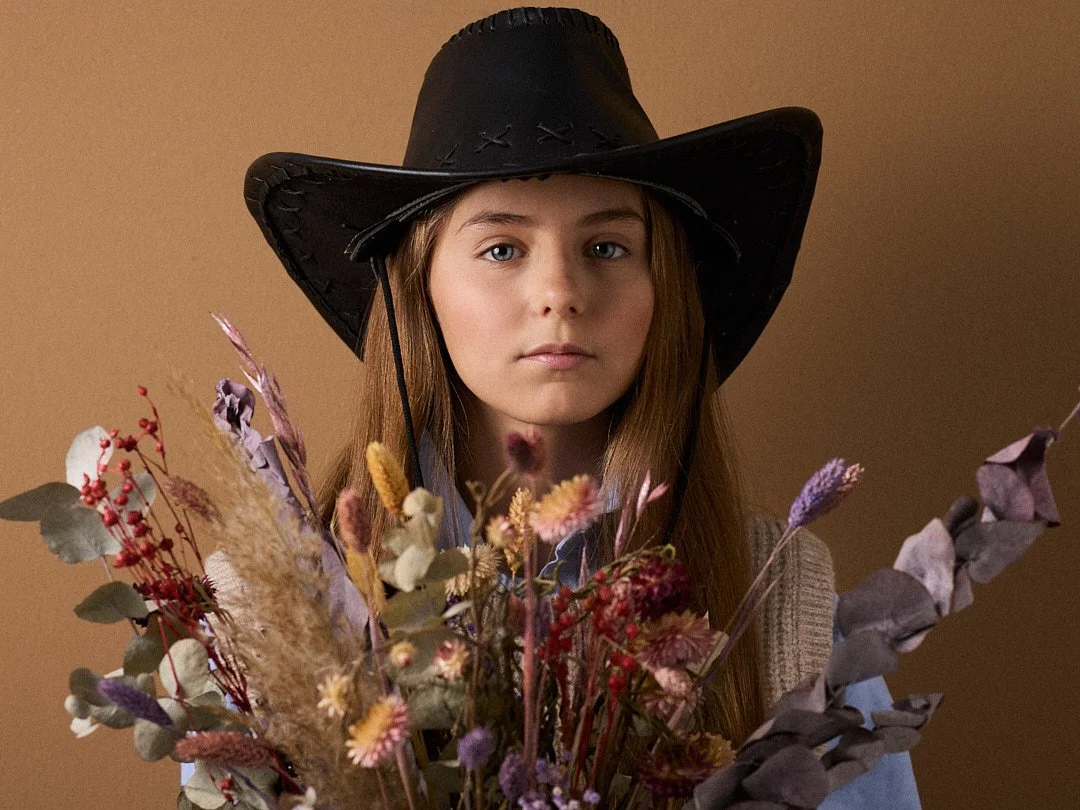 Mädchen mit schwarzem Cowboyhut hinter einem bunten Blumenstrauß, Blick direkt zur Kamera, neutraler Hintergrund.