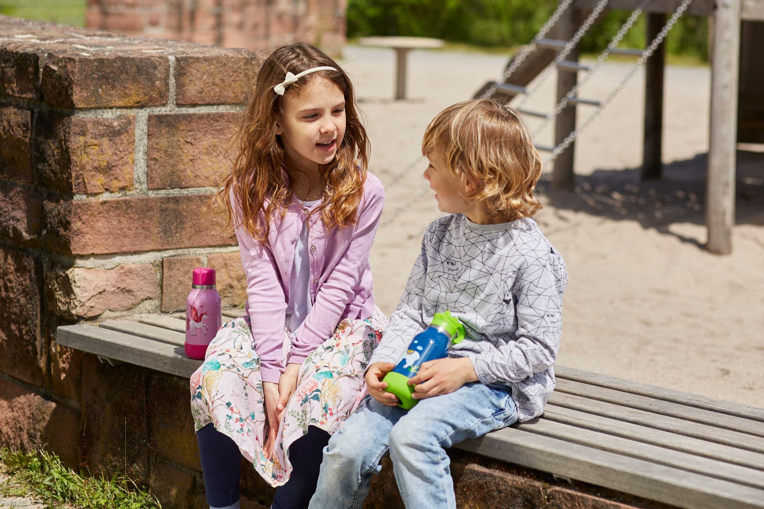 Zwei Kinder sitzen auf einer Bank im Freien, ein Mädchen mit langem, lockigem, braunem Haar und ein Junge mit mittellang, lockigem, blondem Haar, unterhalten sich. Sie haben Trinkflaschen, und im Hintergrund gibt es Spielgeräte.