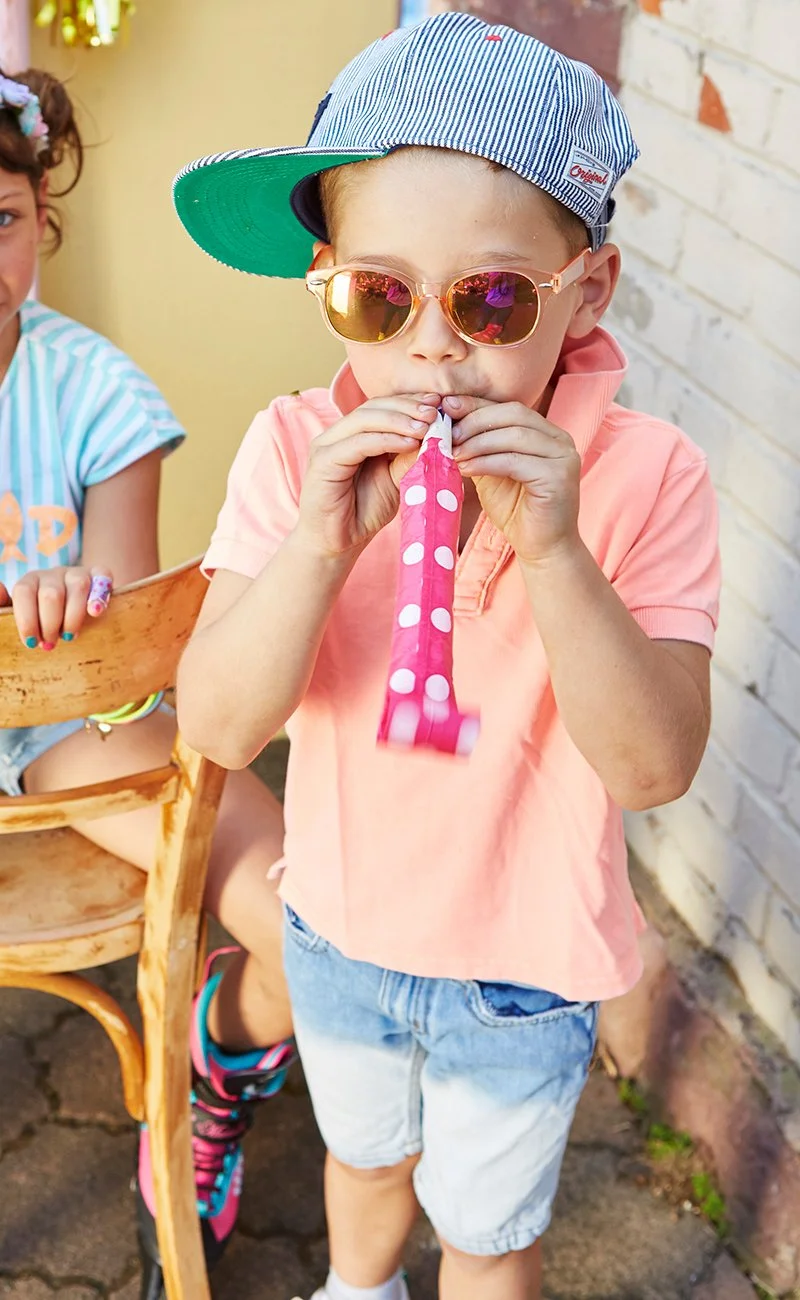 Ein Junge trägt eine rosa Sonnenbrille, eine blaue Kappe, eine rosa Hemd und kurze Hosen. Er bläst auf ein Partyhorn mit rosa und weißen Punkten.