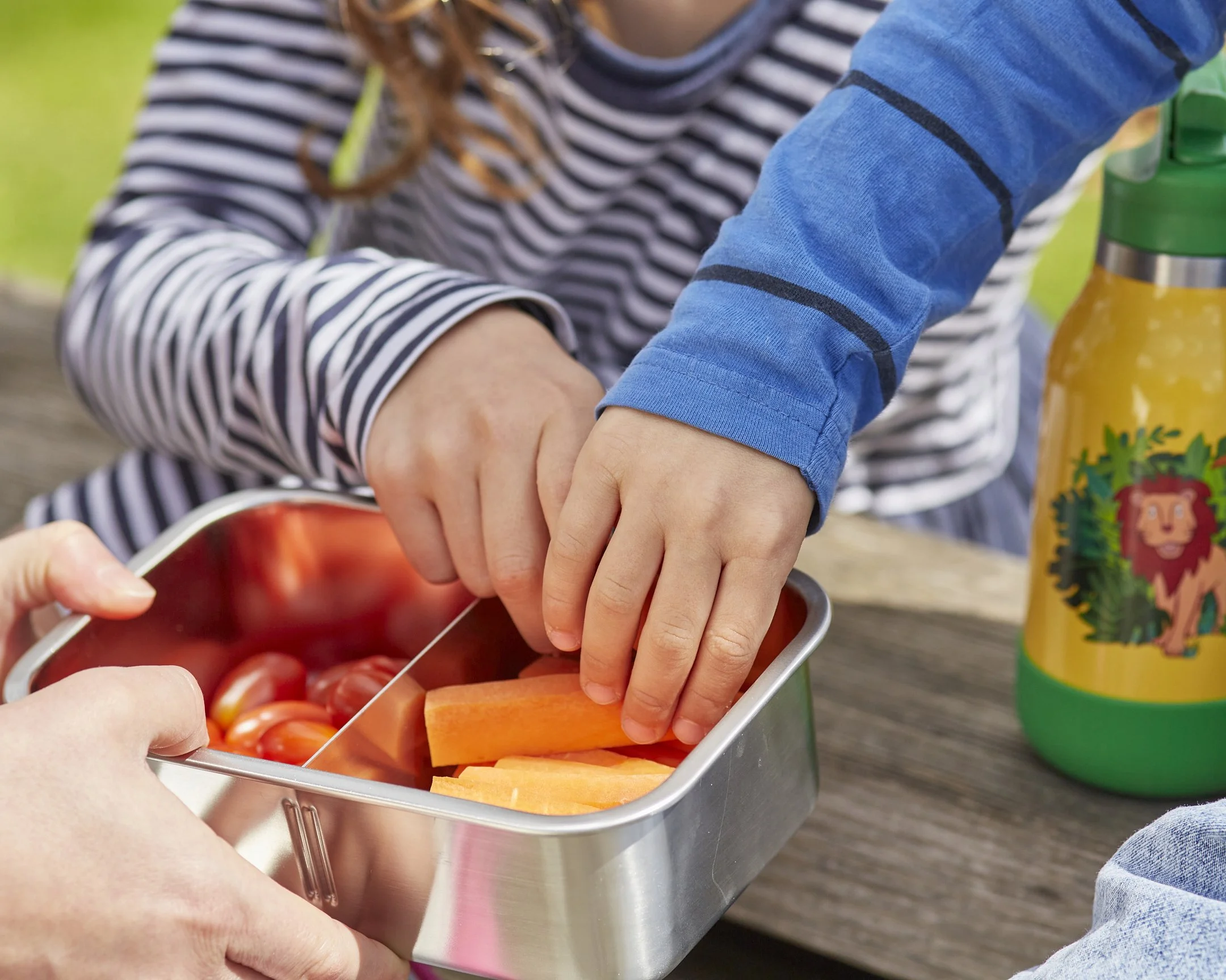 Kinder greifen nach Karotten und Tomaten in einem Metallbehälter im Freien, während eine Trinkflasche mit einem Löwen-Design neben ihnen steht.