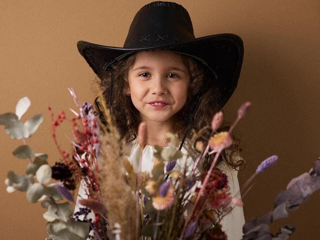 Ein Mädchen mit Locken und einem schwarzen Cowboy-Hut hinter einem Blumenstrauß.