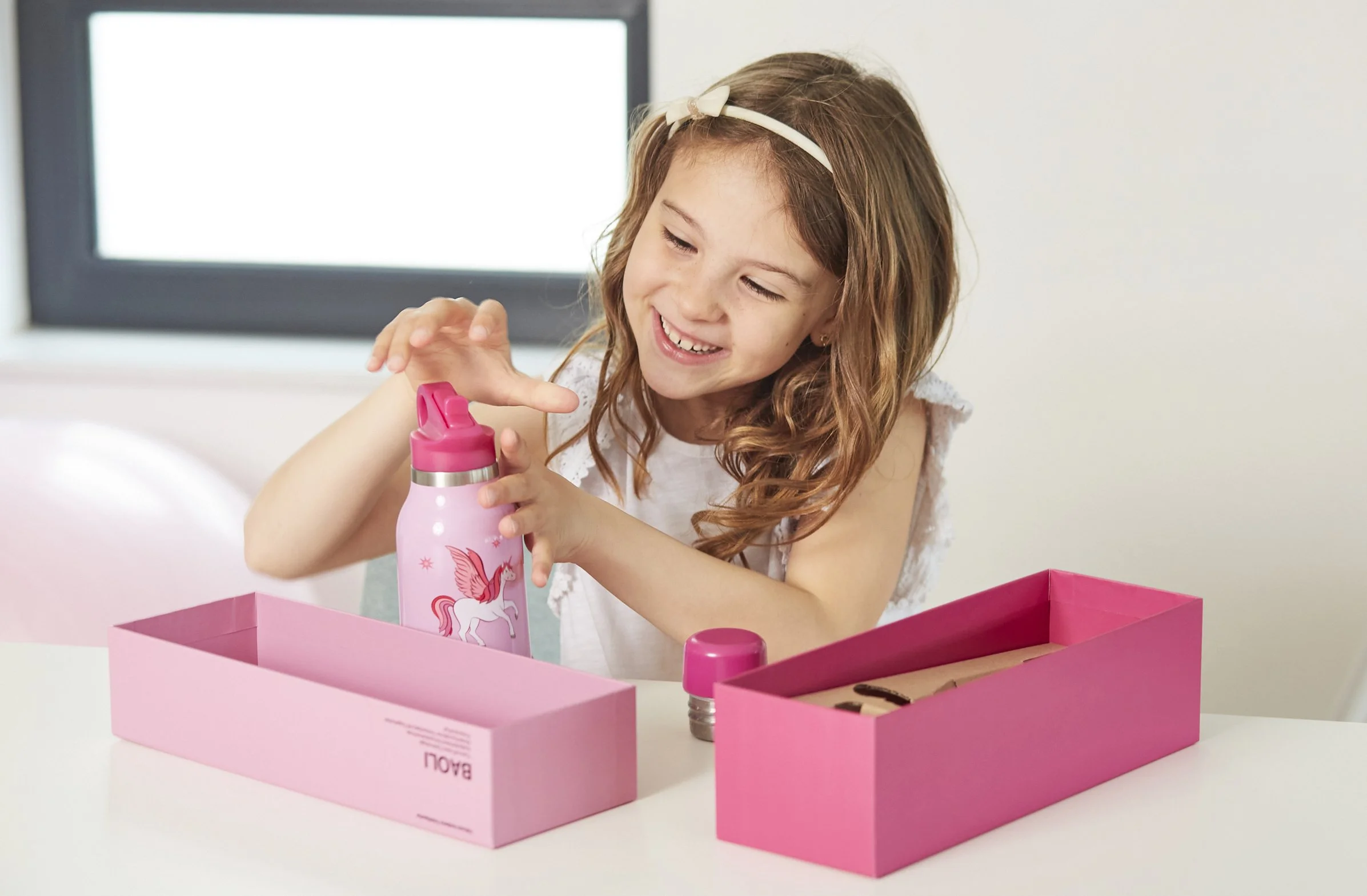Ein lachendes Mädchen mit Langhaar, das einen pinkfarbenen Trinkflasche mit Einhornmotiven in der Hand hält, während sie in einer pinken Schachtel sitzt.