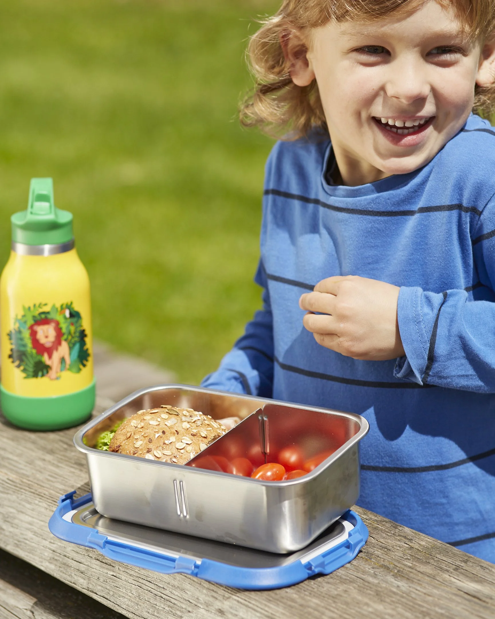 Ein Junge mit blonden Haaren, lachend, trägt ein blaues Hemd mit schwarzen Streifen, sitzt an einem Holztisch im Freien. Auf dem Tisch befindet sich eine gesunde Lunchbox mit einem Vollkornbrötchen und Tomaten, eine Wasserflasche mit Löwenmotiv, und 