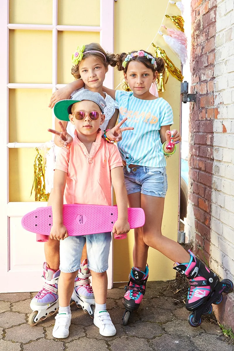 Drei Kinder stehen mit Inlineskates vor einer gelben Tür und einer Backsteinwand, eines hält ein pinkes Skateboard, die Kinder sind verkleidet und feiern vermutlich eine Party.