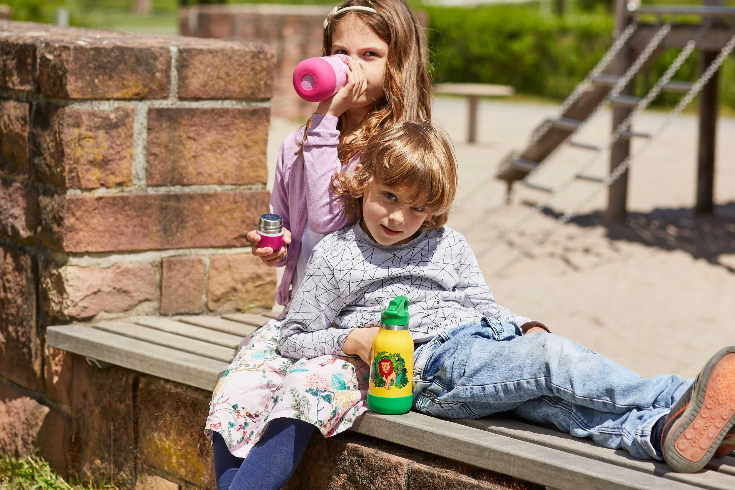 Zwei Kinder, ein Mädchen und ein Junge, sitzen auf einer Holzbank in einem Park. Das Mädchen trinkt aus einer pinkfarbenen Trinkflasche, während der Junge auf der Bank liegt und eine gelbe Trinkflasche mit einem Tierbild neben sich hat.