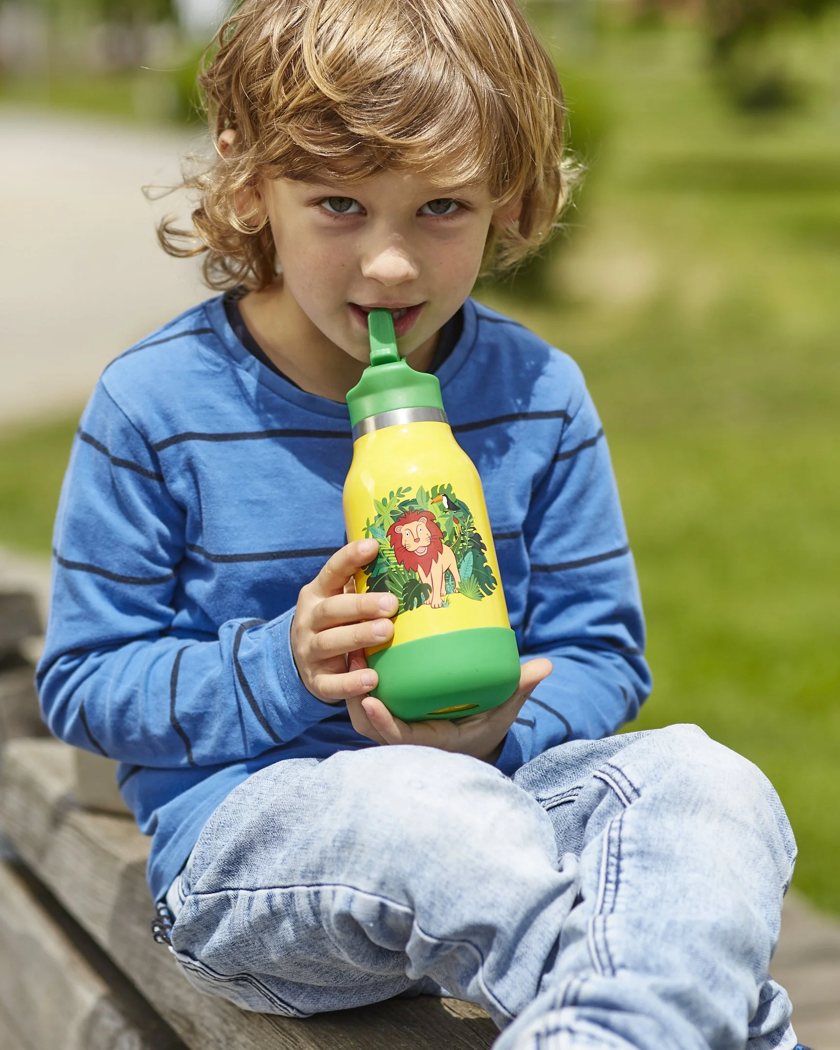 Junge sitzt auf einer Bank im Park und trinkt aus einer gelben Flasche mit Dschungeltiertier-Design, trägt ein blau-gestreiftes Hemd und helle Jeans.