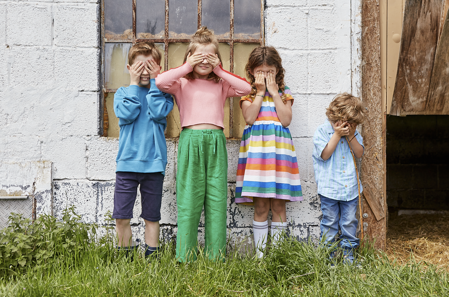 Vier Kinder stehen vor einer weißen Wand neben einem alten Gebäude, alle mit verschlossenen Augen und Händen vor Gesicht, als ob sie nicht hinschauen wollen.