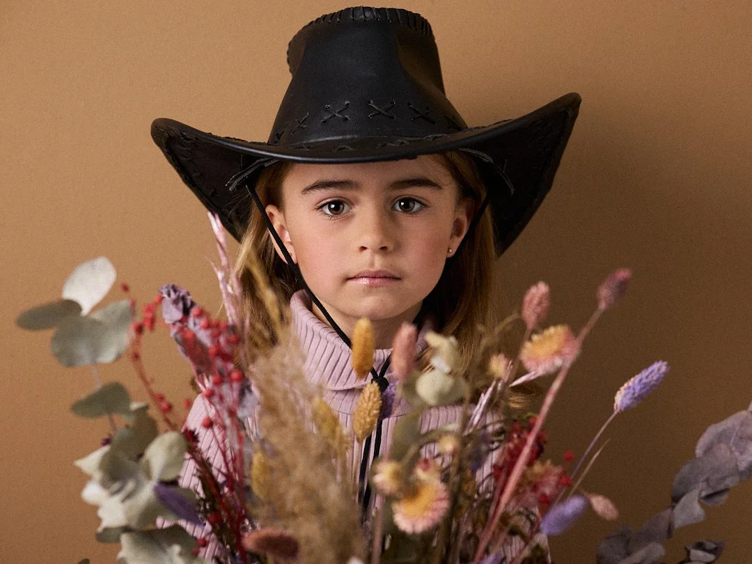 Ein junges Mädchen mit braunen Haaren trägt einen schwarzen Cowboyhut und steht vor einem Buschen von Blumen.