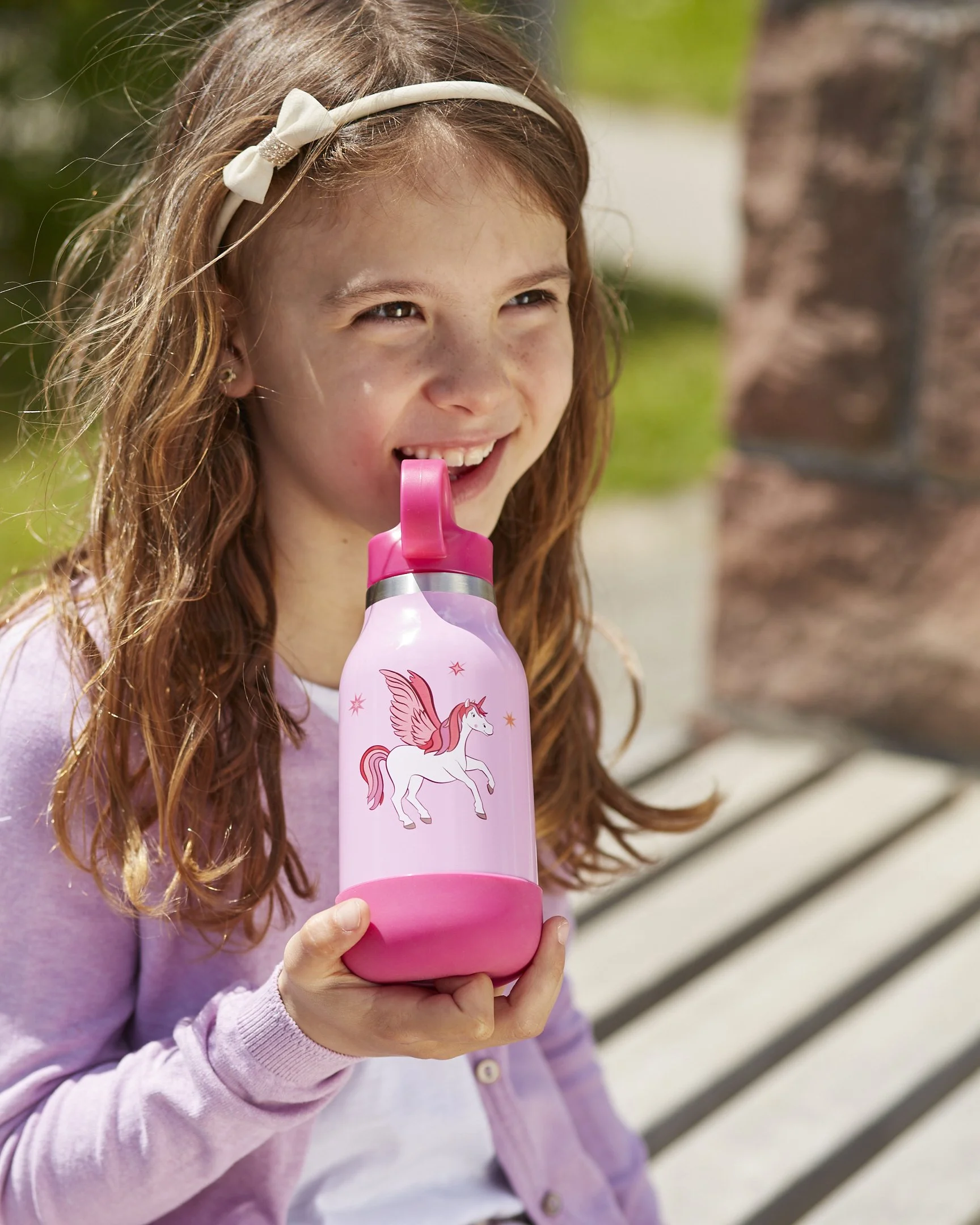 Ein junges Mädchen sitzt auf einer Bank, trinkt aus einer pinken Wasserflasche mit Einhorn-Design, trägt eine weißen Kopfband und ein lila Pullover.