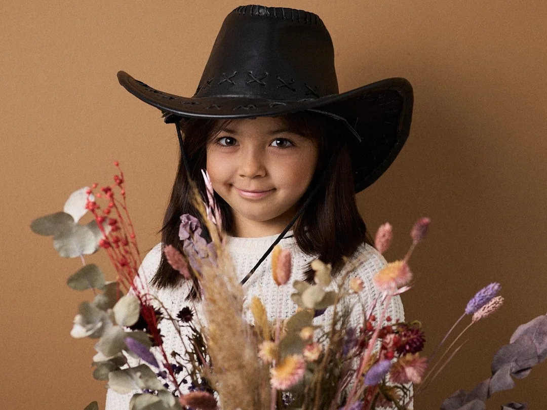Ein junges Mädchen mit braunen Haaren und braunen Augen, trägt einen schwarzen Cowboyhut und ein weißes Pullover, steht vor einem braunen Hintergrund, hält einen Blumenstrauß mit getrockneten Blumen.