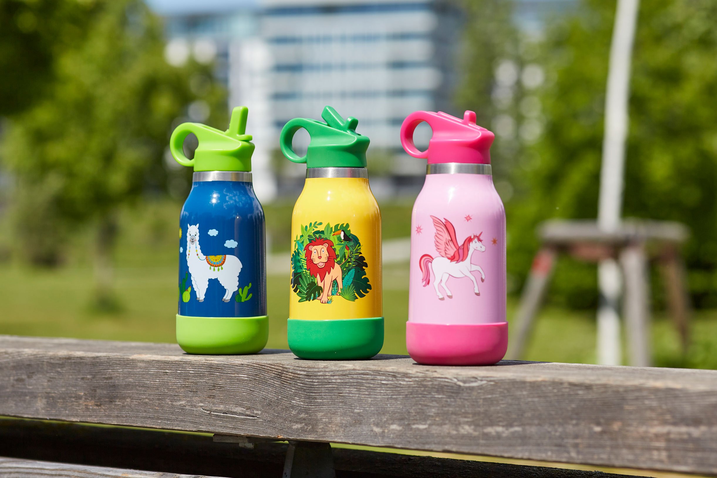 Drei bunte Trinkflaschen mit Tiermotiven auf einer Holzbank im Freien.
