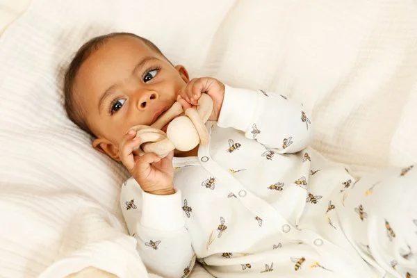 Ein kleiner Junge liegt auf einem weißen Bett und hält einen Beissring aus Holz im Mund, während er bequem in seinen flauschigen Kissen liegt.