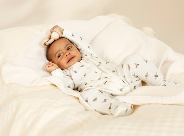 Ein Baby liegt auf einem Bett in einem weißen Schlafanzug mit Tiermotiven, mit einer Plüschfigur auf dem Kopf, und schaut in die Kamera.