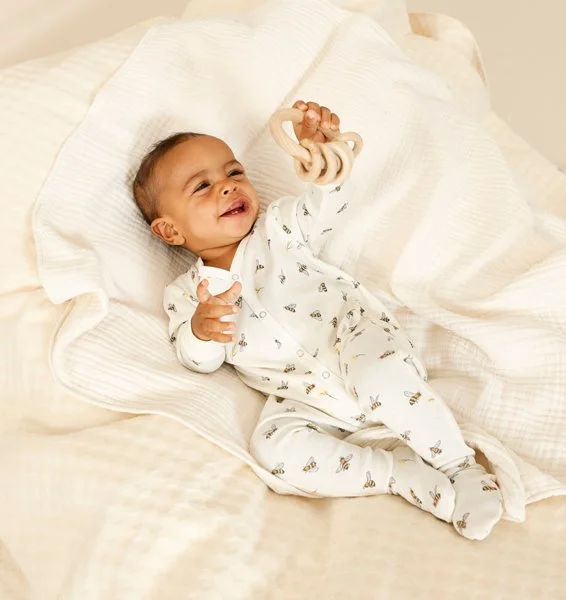 Ein lachendes Baby liegt auf einem Bett in weißen pyjamaartigen Kleidungsstücken, hält einen Schnuller in der Hand und sieht fröhlich aus.