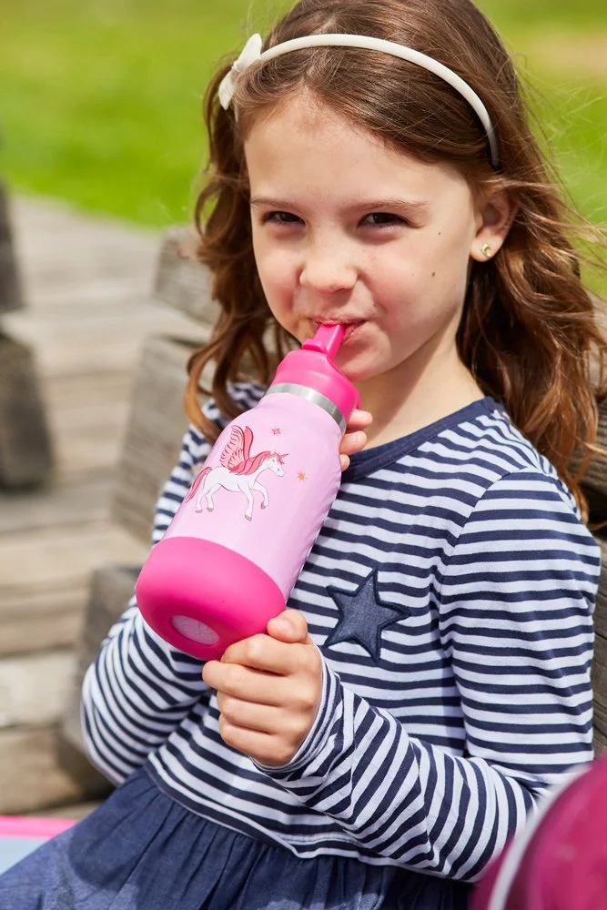 Ein Mädchen mit braunen, lockigen Haaren und Schleierkopfband trinkt aus einer rosa Wasserflasche mit Einhorn-Design im Freien.