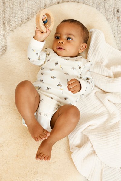 Ein Baby liegt auf einem weißen Kissen und hält ein Holzspielzeug in der Hand.