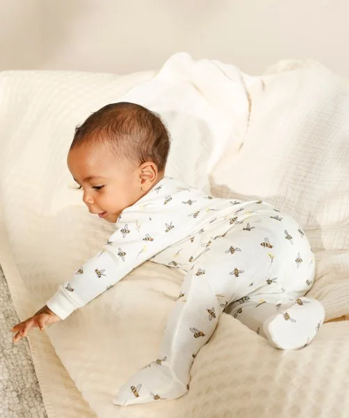 Kleines Baby in weißem Schlafanzug mit Bienenmuster, das auf einem weißen Sofa, krabbelnd, sitzt.