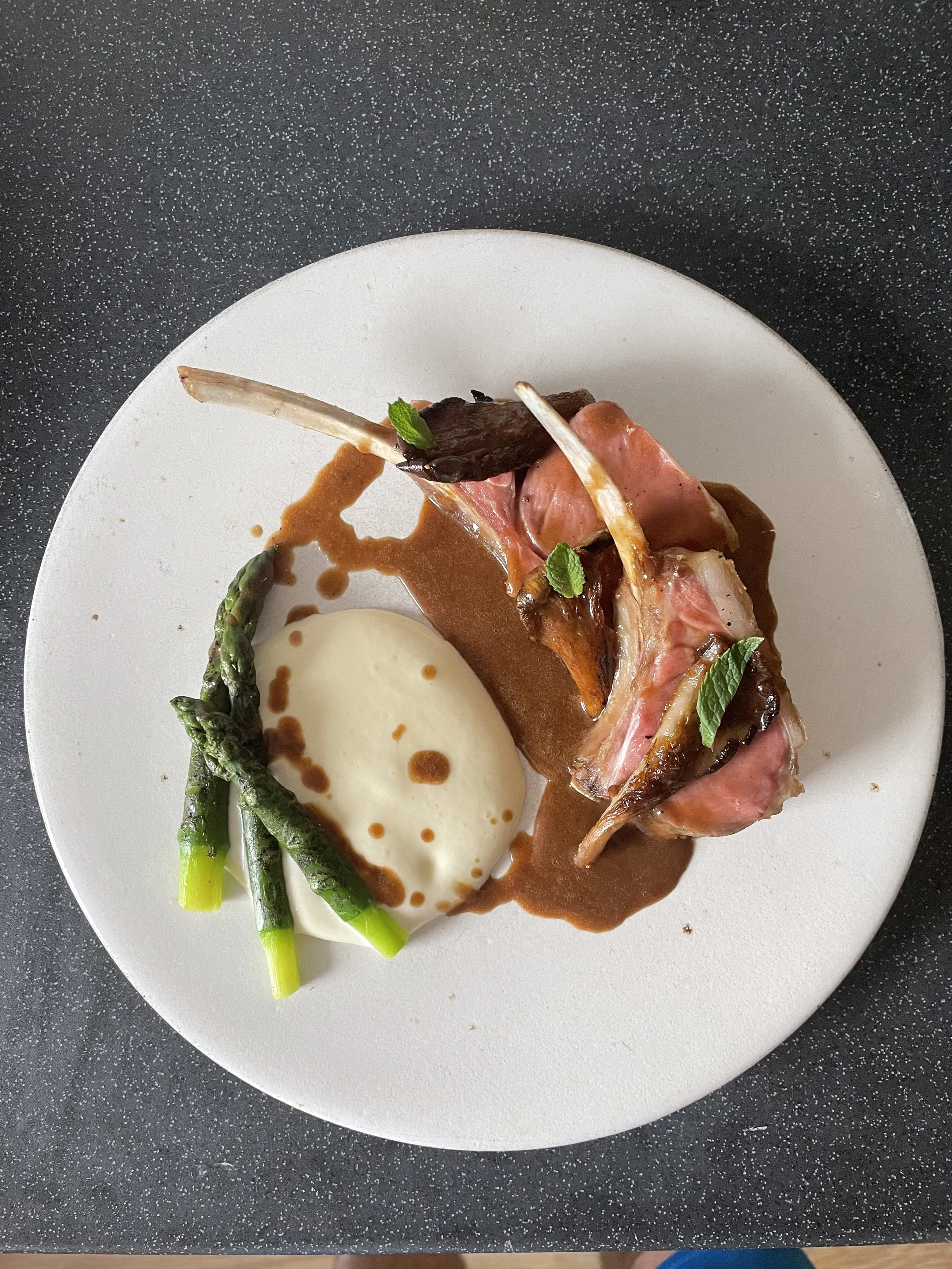 A plated gourmet dish featuring a portion of lamb with a bone, garnished with herbs, accompanied by green asparagus and a dollop of creamy mashed potatoes with brown sauce.