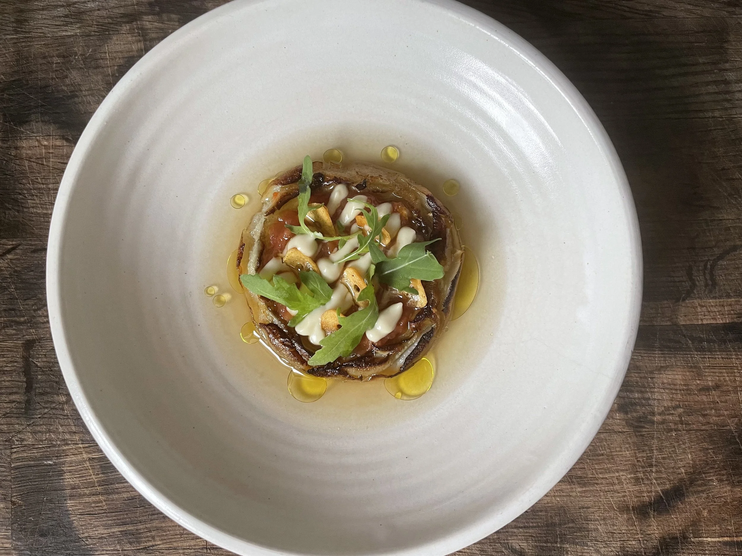 A plated dish with grilled vegetables topped with microgreens and a drizzle of oil, served on a white plate on a wooden surface.