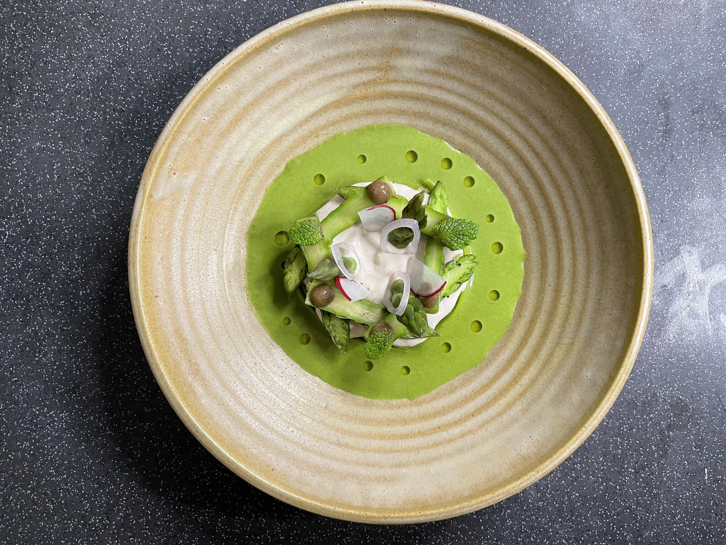 A ceramic bowl with a green veloute with a dollop of cream and a small salad of asparagus, radish slices, and mint leaves in the center.