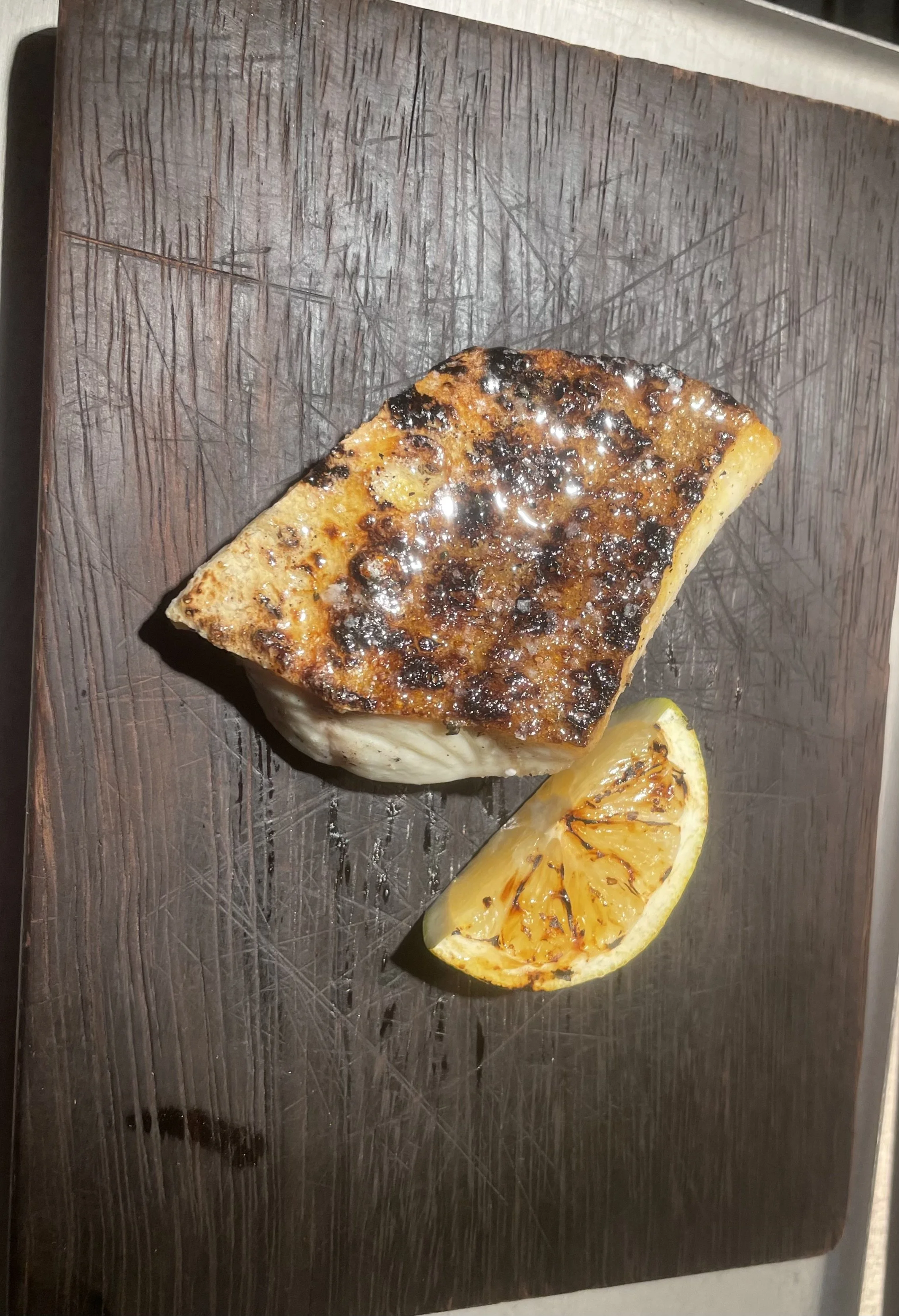 Grilled piece of fish with a lemon wedge on a dark wooden cutting board.