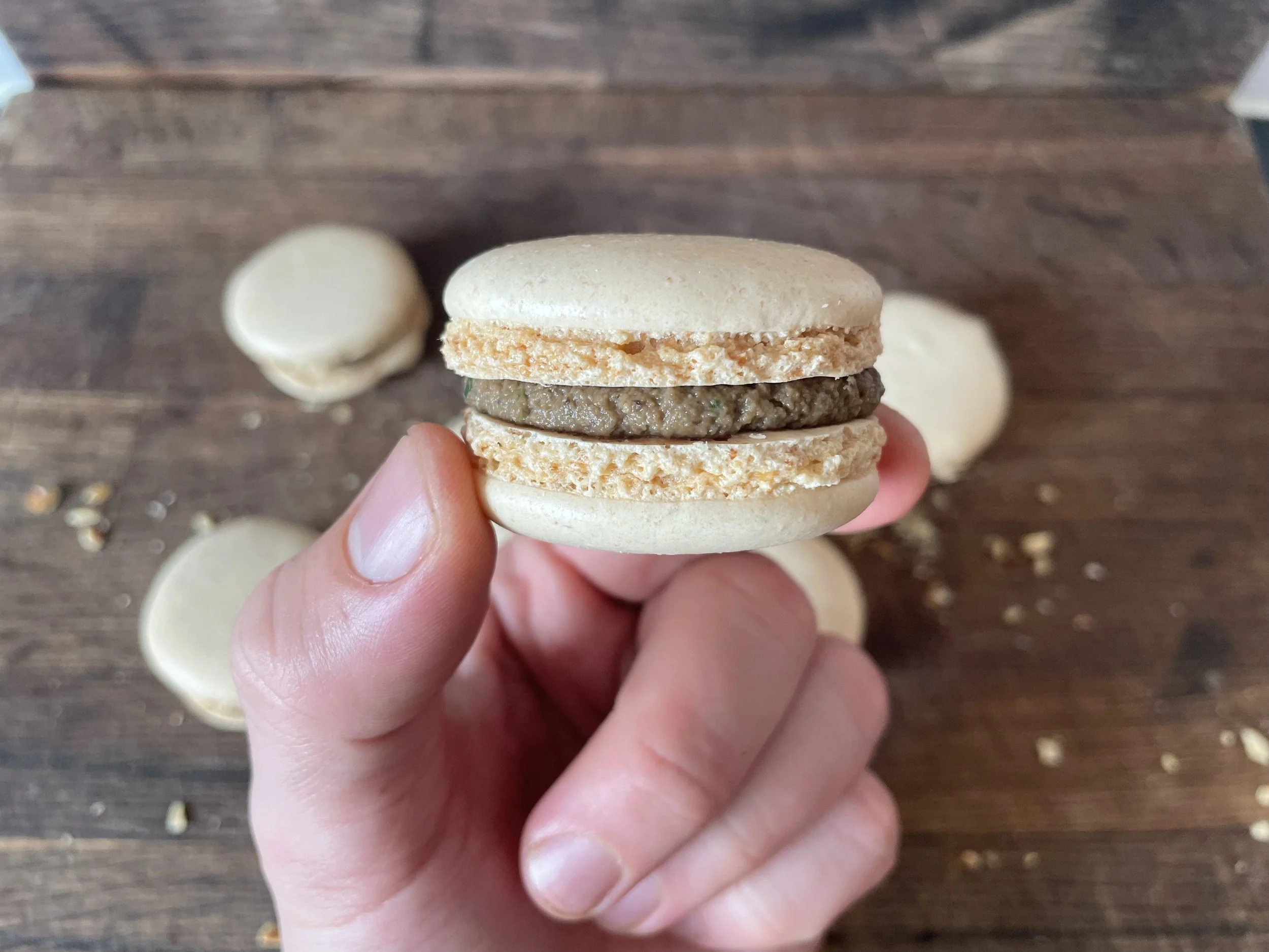 Hand holding a beige macaroon with a chocolate filling, three more macarons on a wooden surface in the background, with crumbs scattered around.