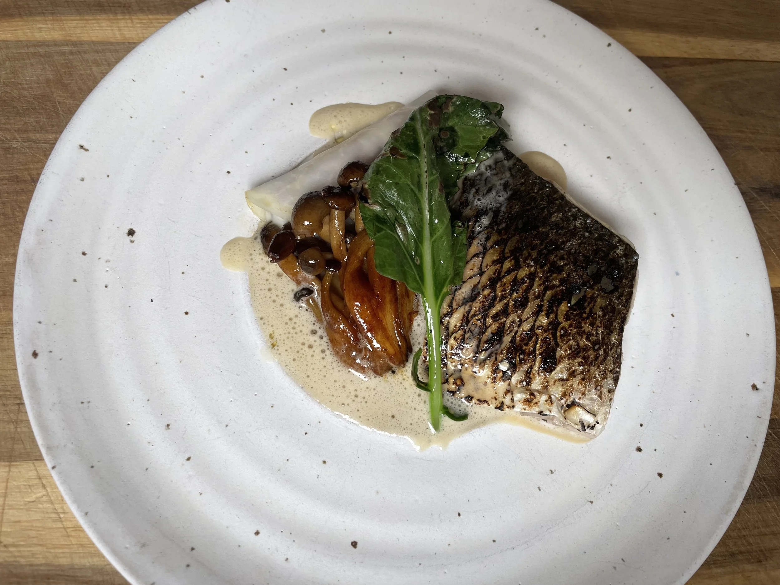 Pan-seared fish fillet garnished with a green leafy herb, served with sautéed mushrooms and caramelized onions, and a light cream sauce on a white plate.
