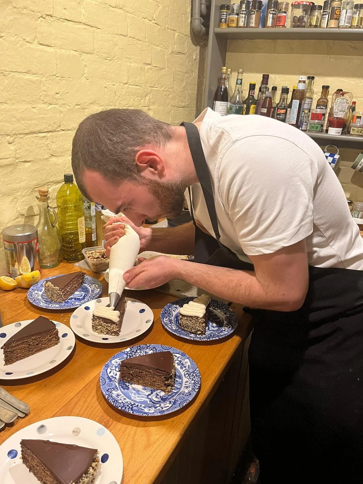 A man in a white shirt and black apron decorates slices of layered chocolate cake with white icing at a wooden table.