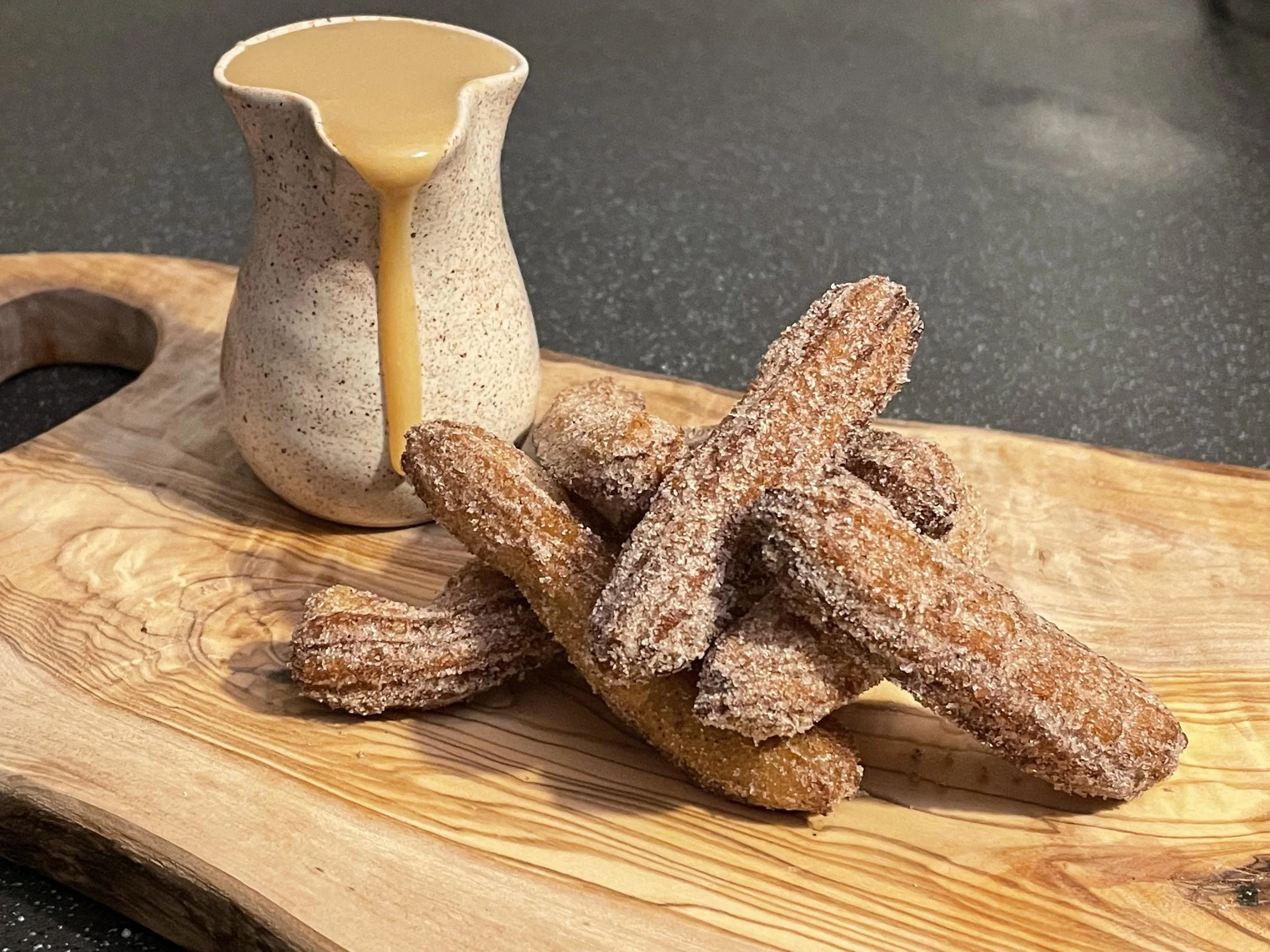 Fried churros coated in sugar on a wooden serving board, with a small pitcher of caramel sauce nearby.