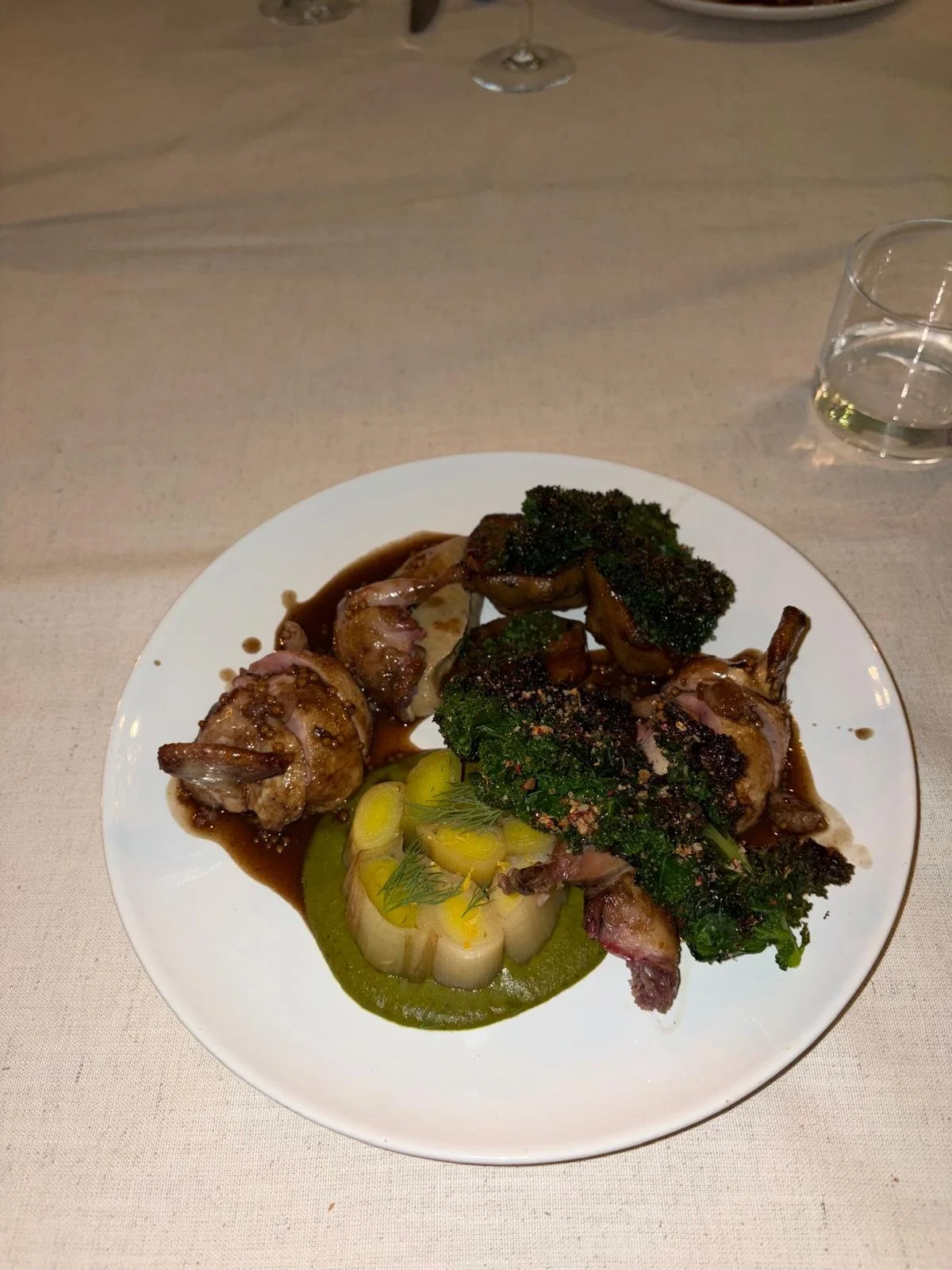 A plated dish with roasted chicken, broccoli, and a vegetable garnish, served with a dark sauce on a white plate. A wine glass and a water glass are visible in the background.
