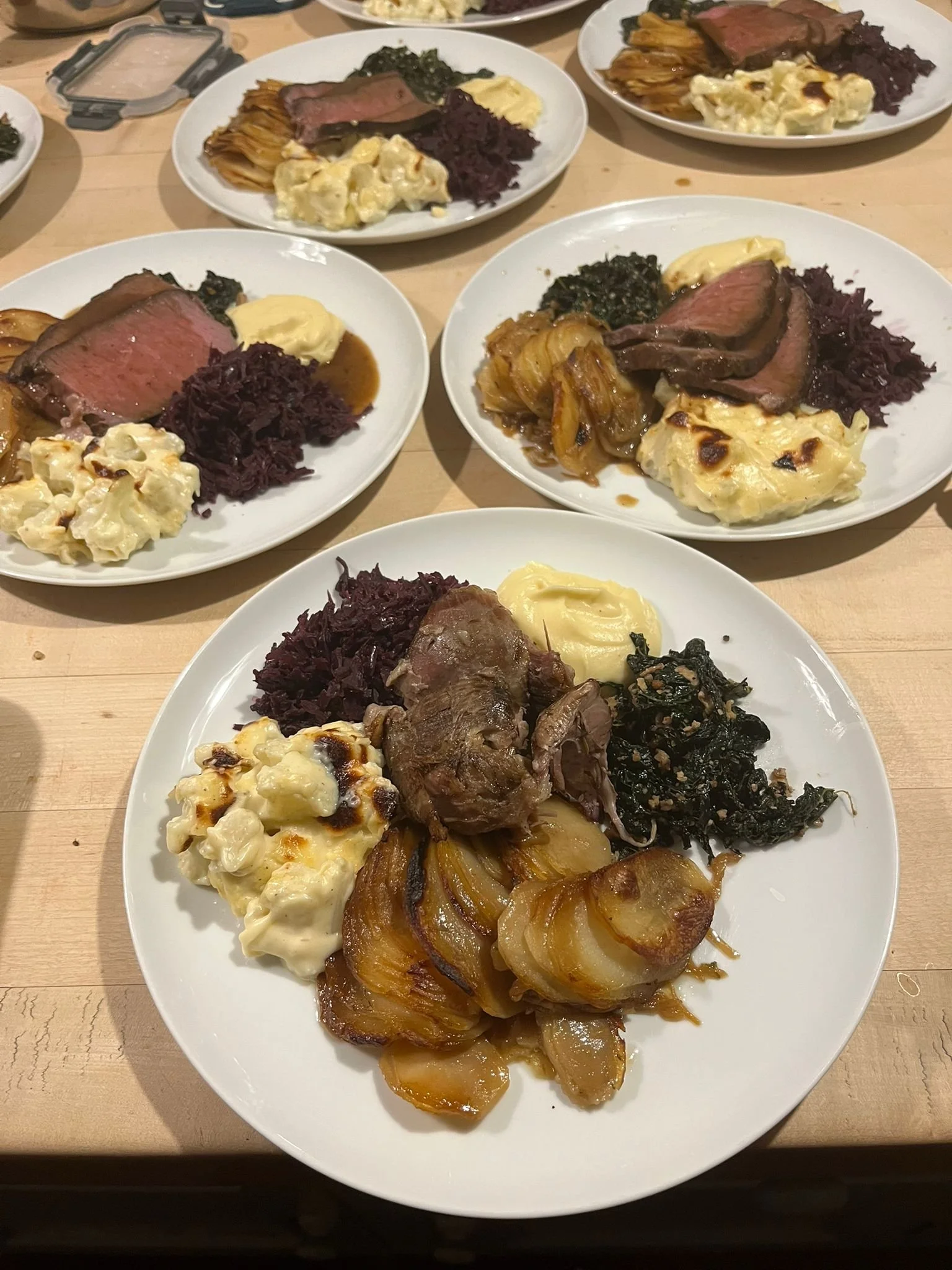 Plates of food including roast beef, mashed potatoes, caramelized onions, sautéed greens, and red cabbage on a wooden table.