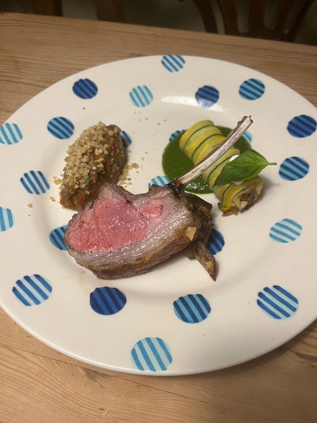A plated serving of medium-rare lamb chop with a breadcrumb crust, a quenelle of vegetable pureé, and a rolled vegetable wrap garnished with a basil leaf.