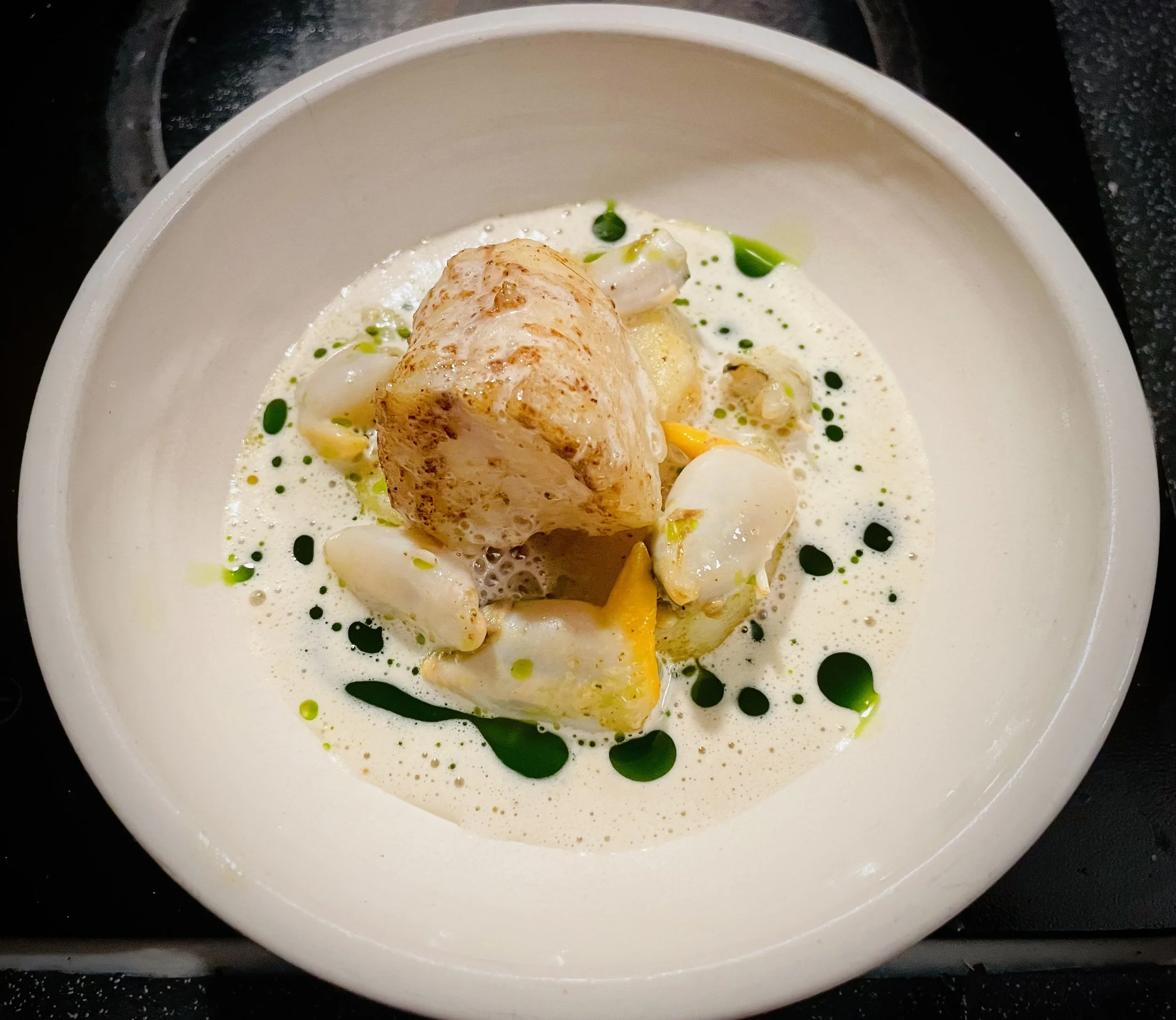 A dish with a large, seared piece of fish served on a bed of foam, garnished with chopped vegetables and green oil drops.